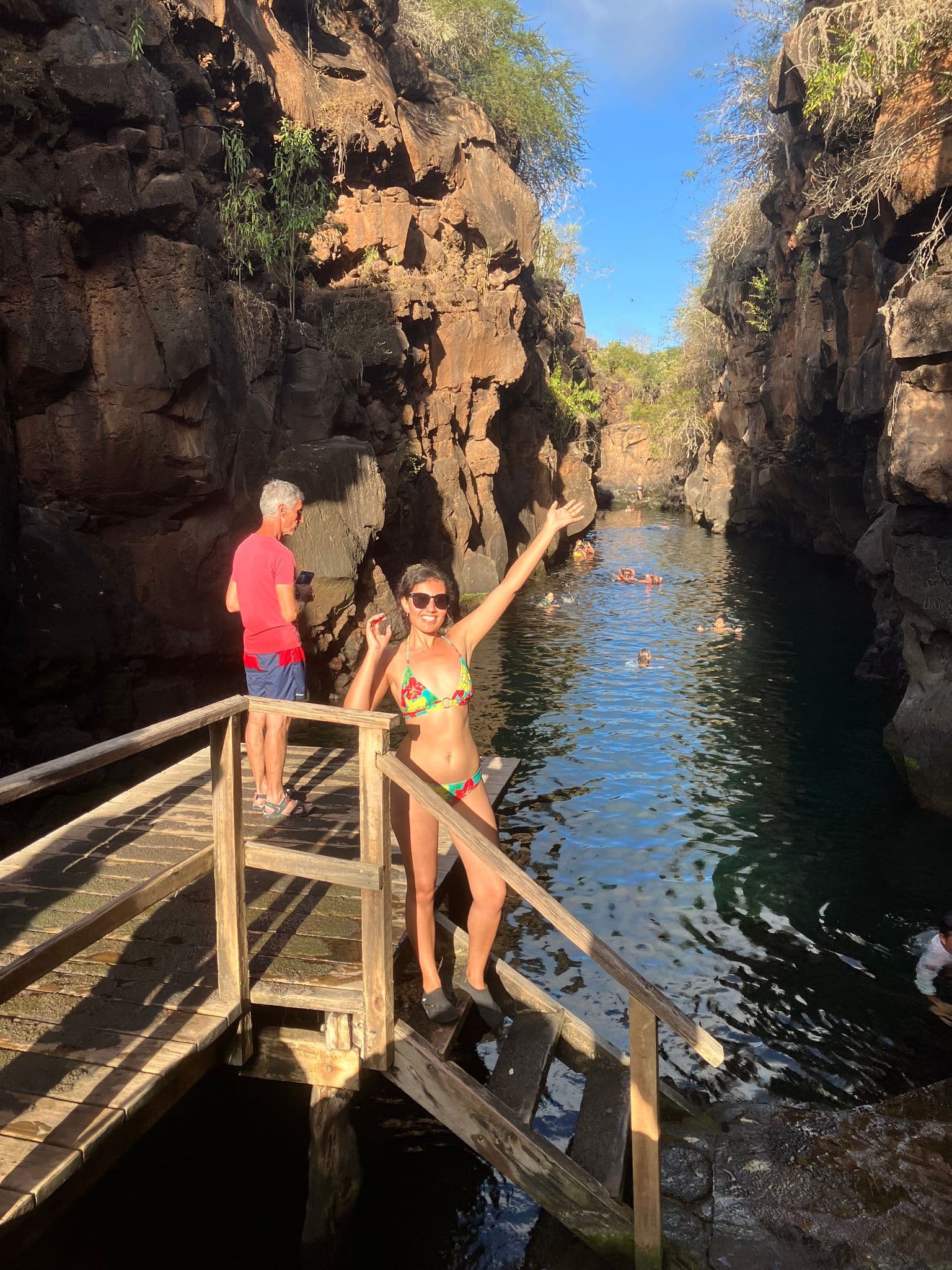 Volcanic rock channel in the Galápagos Islands with a woman waving from wooden steps and swimmers in the water, Ecuador.