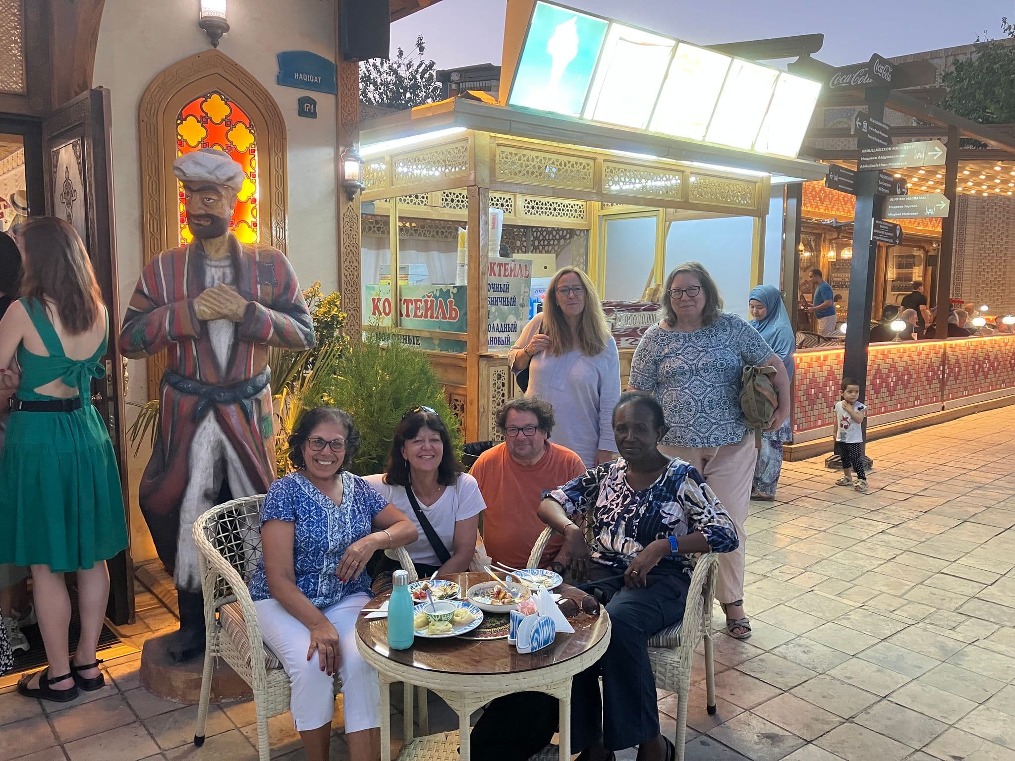 Group of travelers sitting at a table with plates in a lively outdoor bazaar near a decorative stall in Uzbekistan.