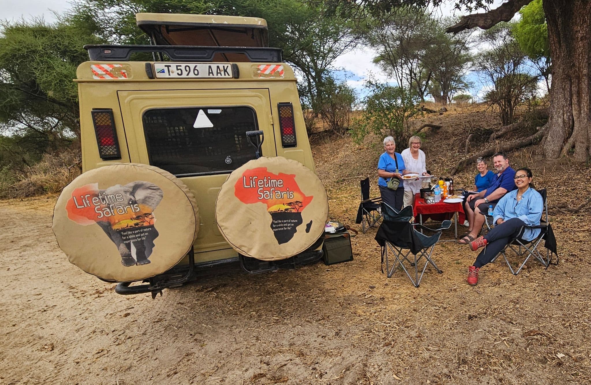 Travelers having an outdoor lunch beside a safari vehicle under acacia trees in Tarangire National Park, Tanzania.