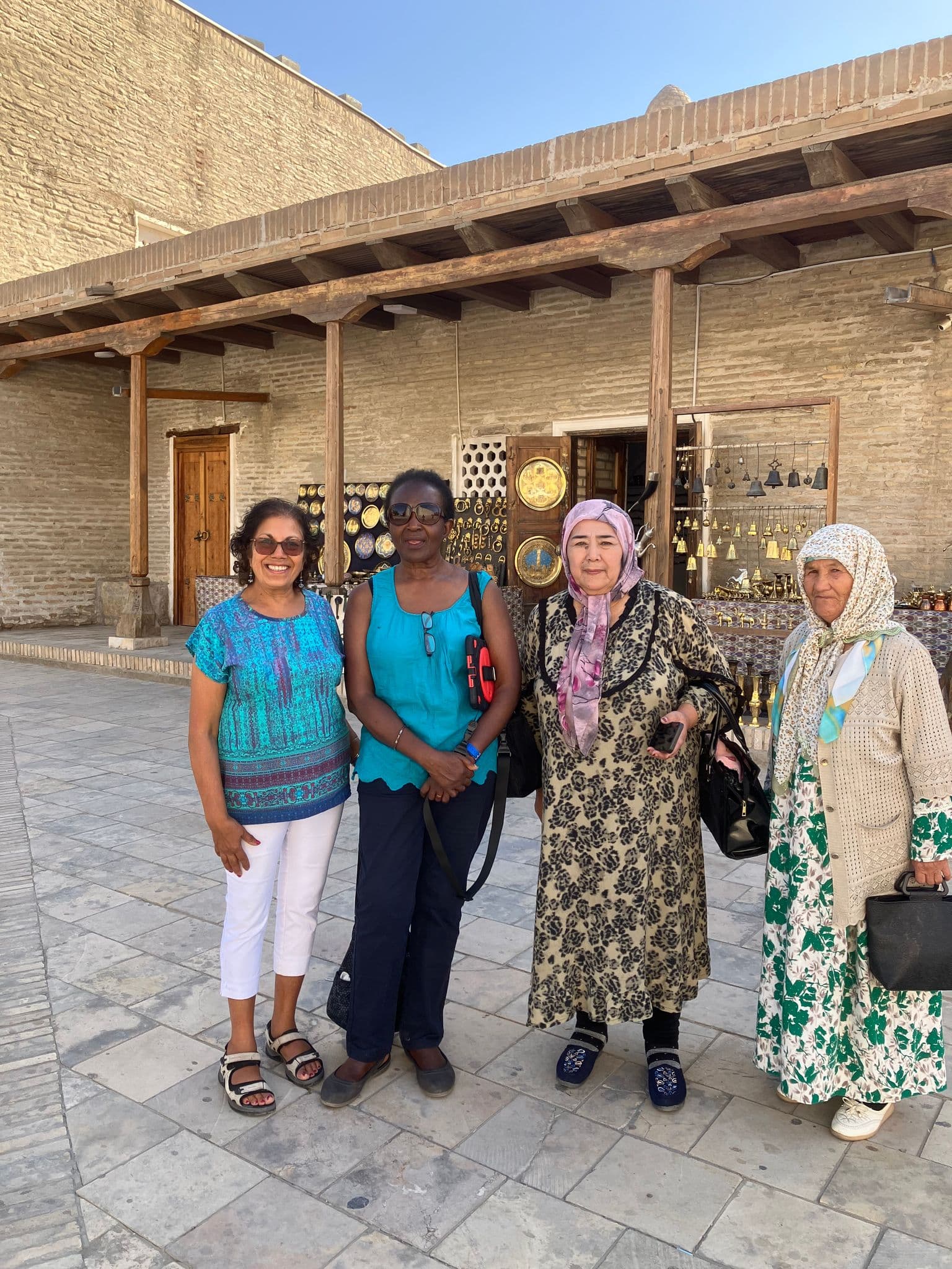 Four women posing in front of brassware and market stalls in a sunlit brick courtyard in Uzbekistan.