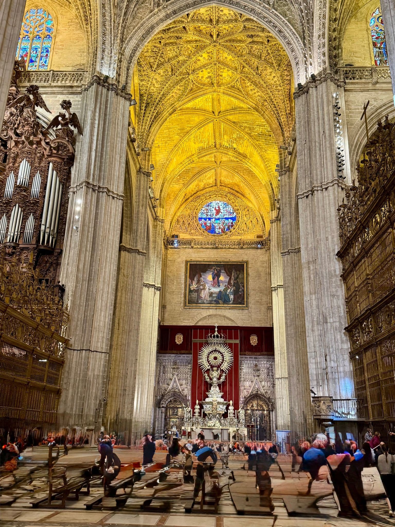 Seville Cathedral interior with gilded vaulted ceiling and ornate altar, visitors seated and walking on benches, Seville, Spain.