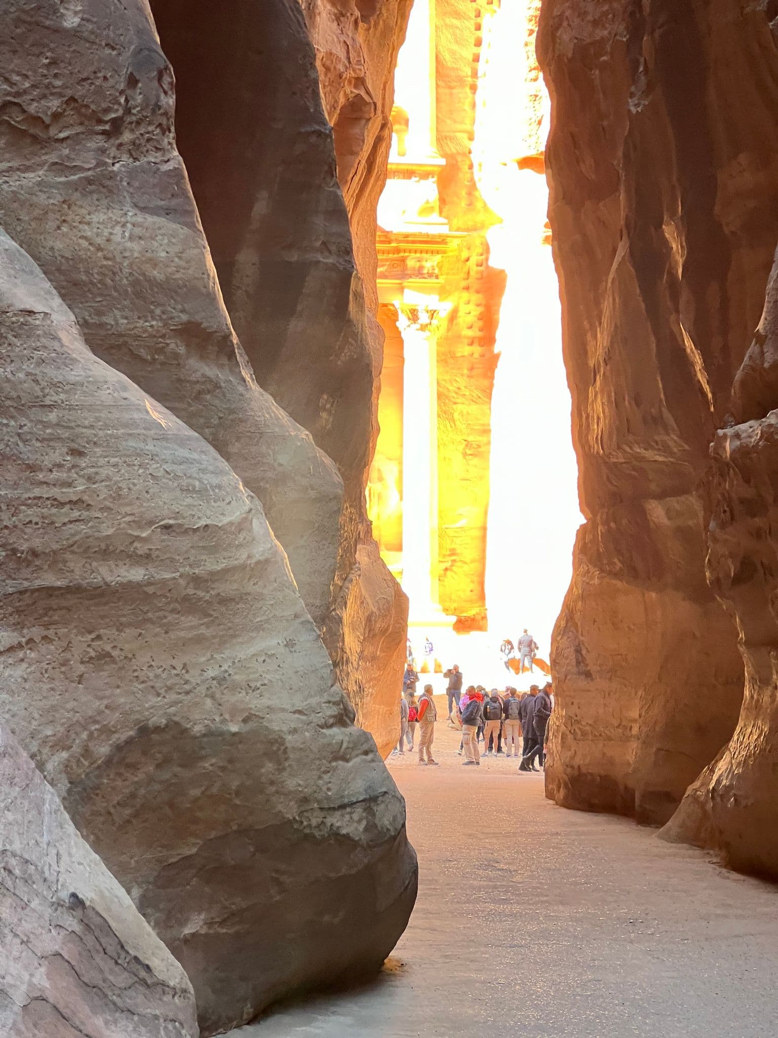 Al-Khazneh (the Treasury) seen through the Siq with tourists on the sandy path as morning sun lights the sandstone, Petra, Jordan.