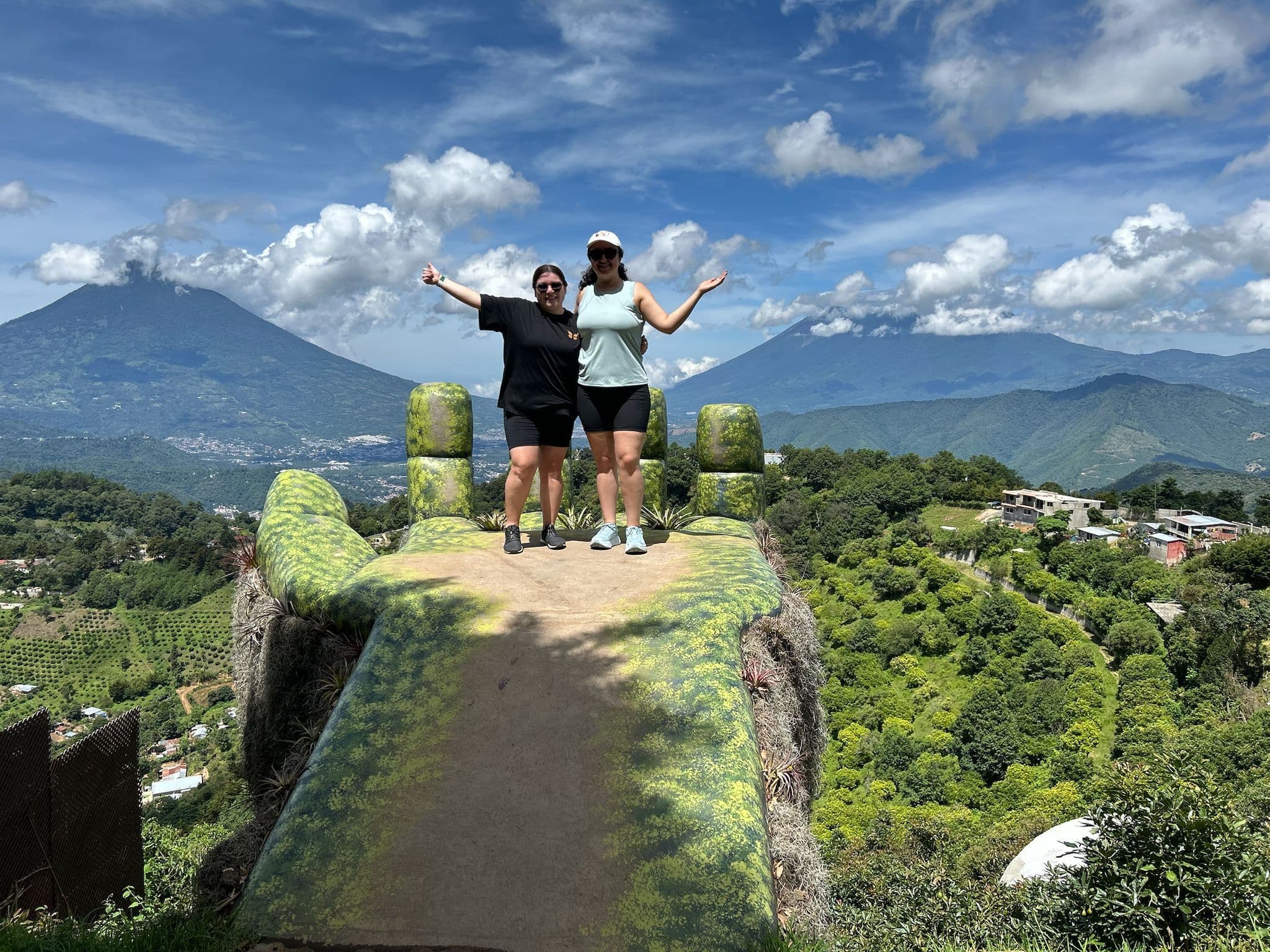 Hand-shaped viewpoint at Hobbitenango with two people standing with arms outstretched and volcano peaks behind, Antigua, Guatemala.