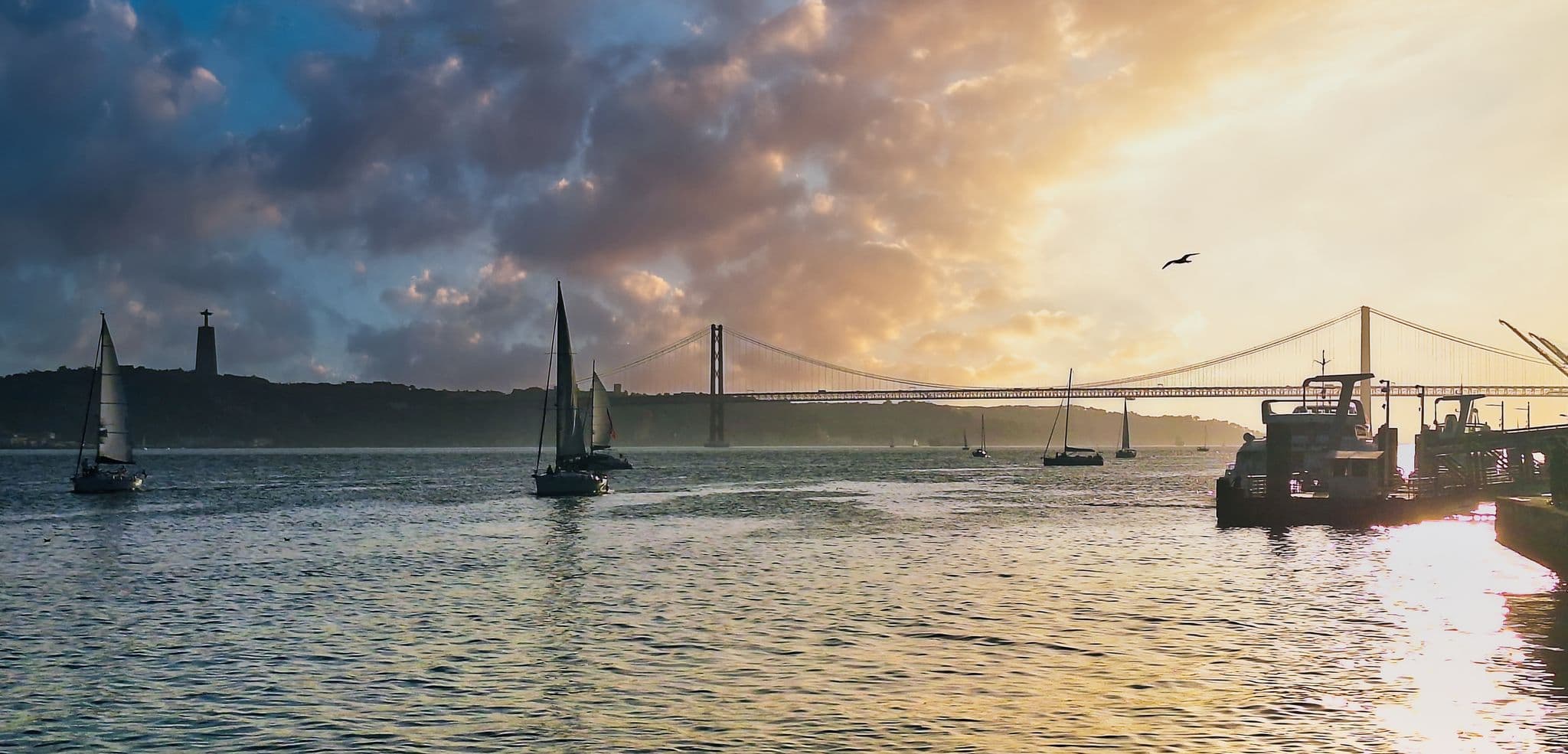 25 de Abril Bridge and the Cristo Rei statue over the Tagus River with sailboats at sunset in Lisbon, Portugal.