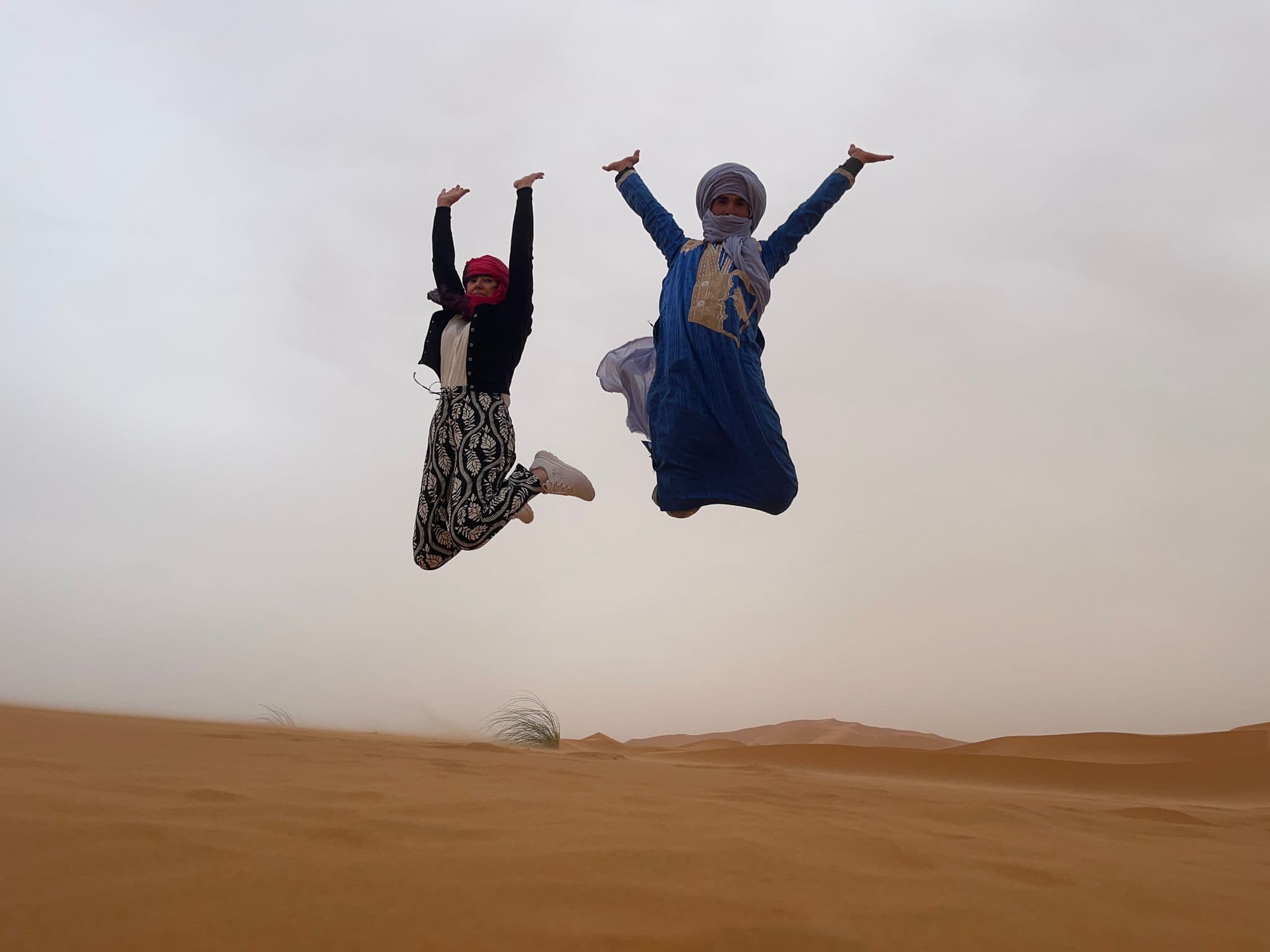 Erg Chebbi sand dunes with two people mid-jump, arms raised, near Merzouga, Morocco.