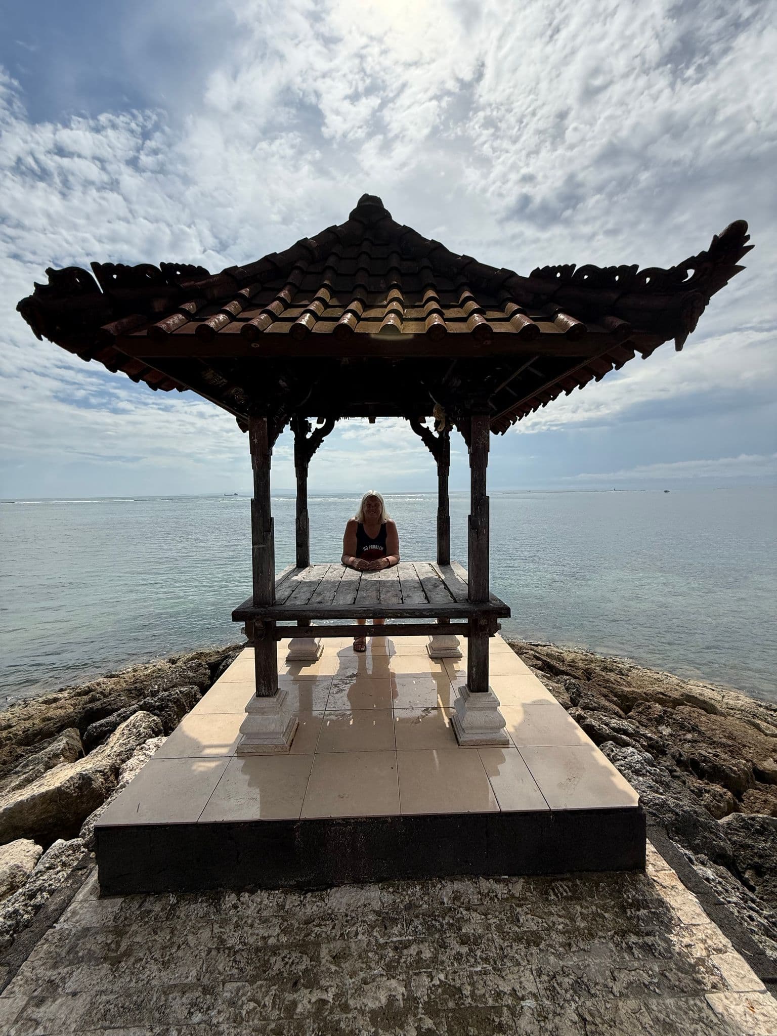 Balinese wooden gazebo on a rocky shore with a person sitting inside, overlooking the sea at Benoa Beach, Bali, Indonesia.
