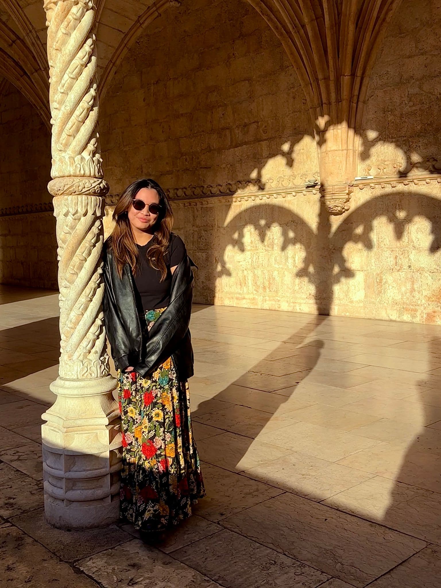 Jerónimos Monastery cloister in Lisbon with a person leaning against a carved column and sunlit arch shadows on the stone wall.