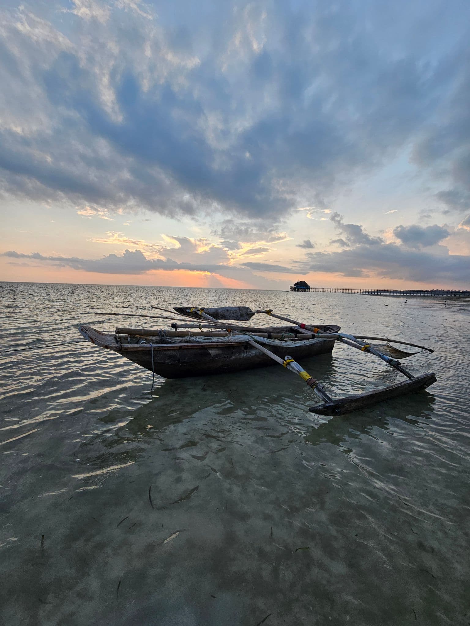 An outrigger canoe resting in shallow water at sunrise off Zanzibar, Tanzania, with a distant pier and pastel sky.