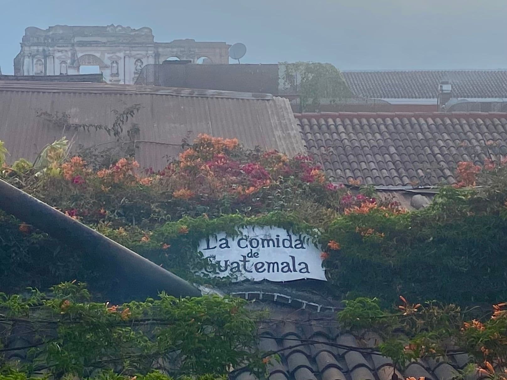 Catedral de Santiago ruins and tiled rooftops framed by flowering vines above a restaurant sign in Antigua Guatemala, Guatemala.