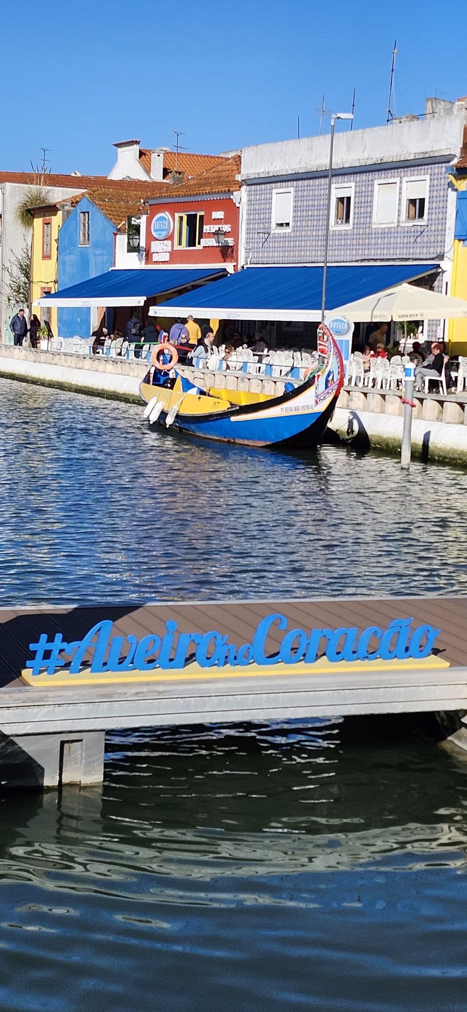 A moliceiro boat docked on the Ria de Aveiro canal in front of colorful waterfront buildings and a blue '#AveironoCoração' sign, Aveiro, Portugal.