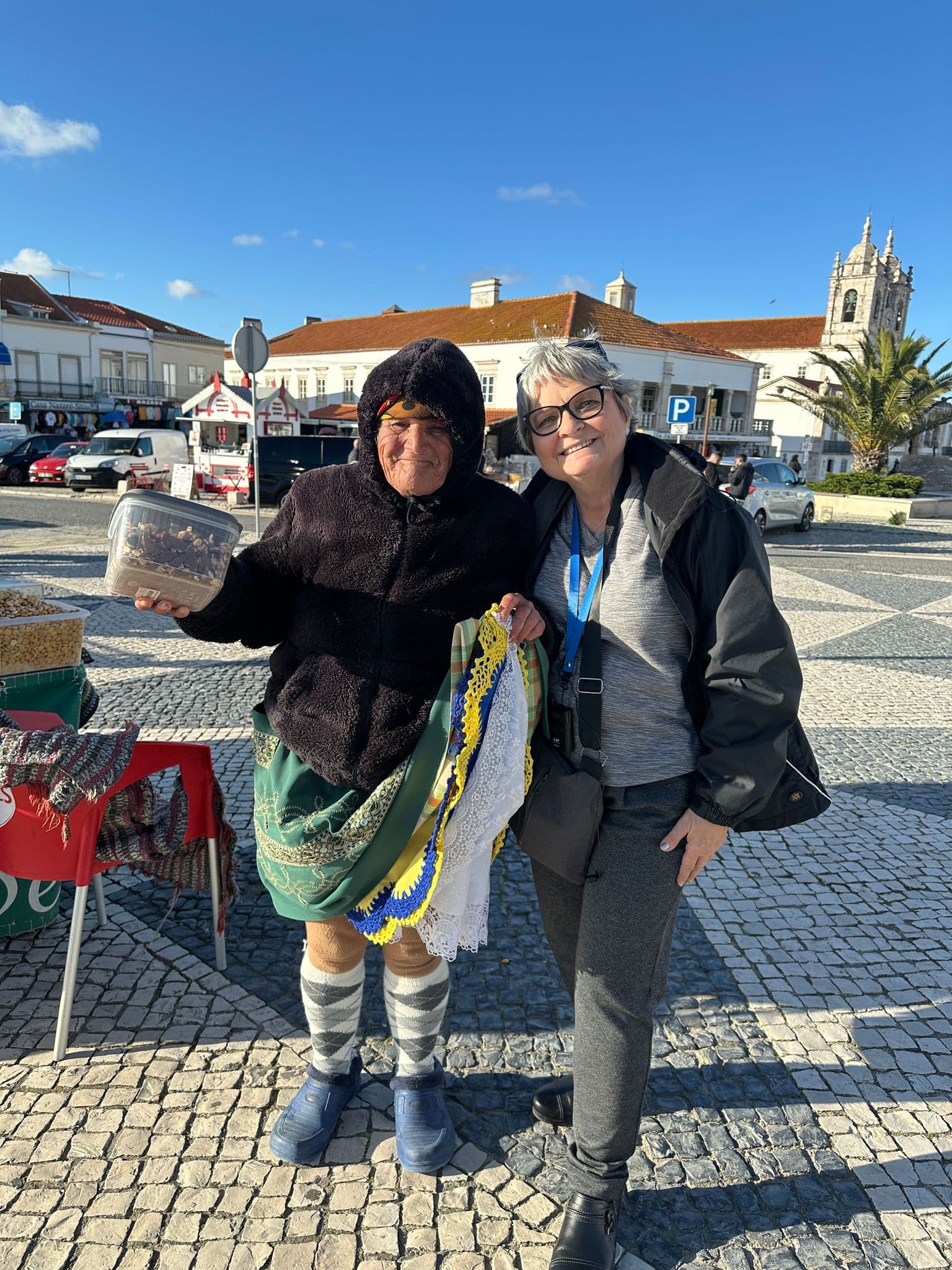 Two women — an elderly "seven skirts" vendor and a tourist — posing in a sunlit square in Nazaré, Portugal.