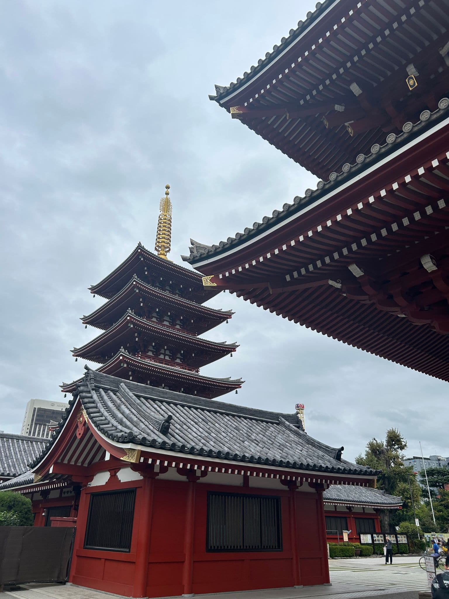 Five-story pagoda and red temple roofs at Senso-ji in Asakusa, Tokyo, Japan, framed against a cloudy sky with a few visitors nearby.