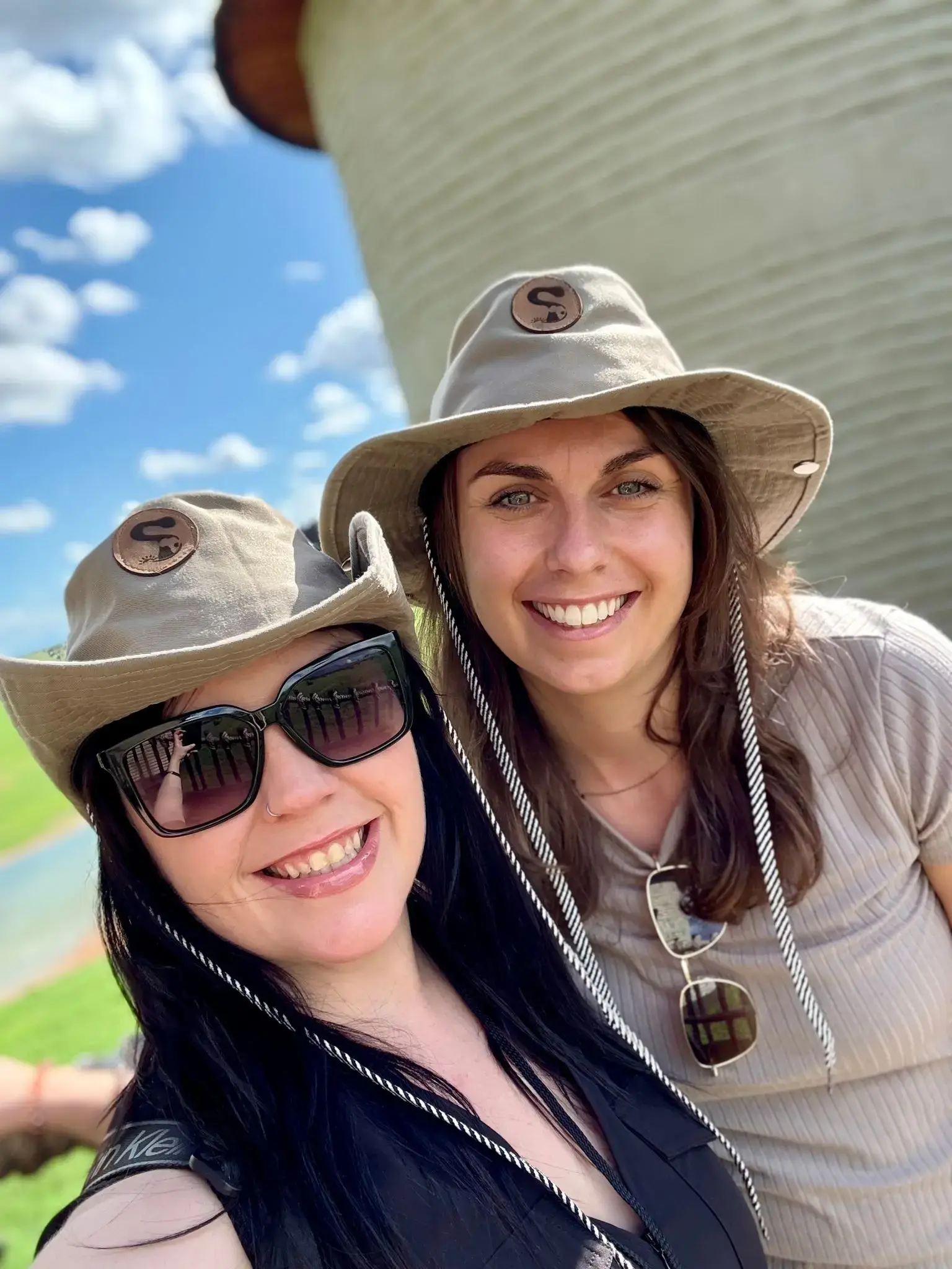 Two women wearing wide-brim hats smiling for a selfie outdoors near water and green fields on a sunny trip.