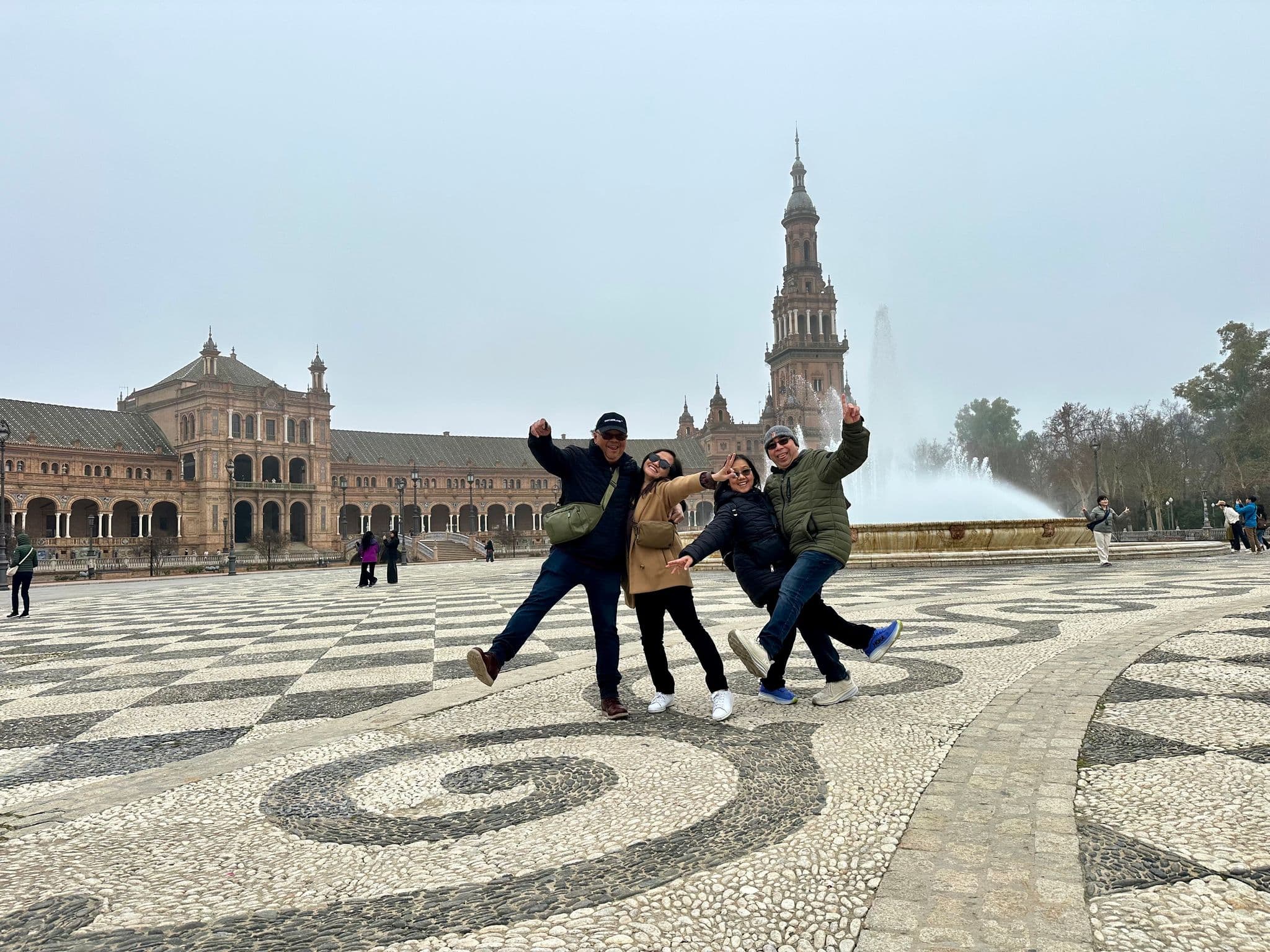 Plaza de España and its central fountain with four travelers posing and kicking on the patterned plaza tiles, Seville, Spain
