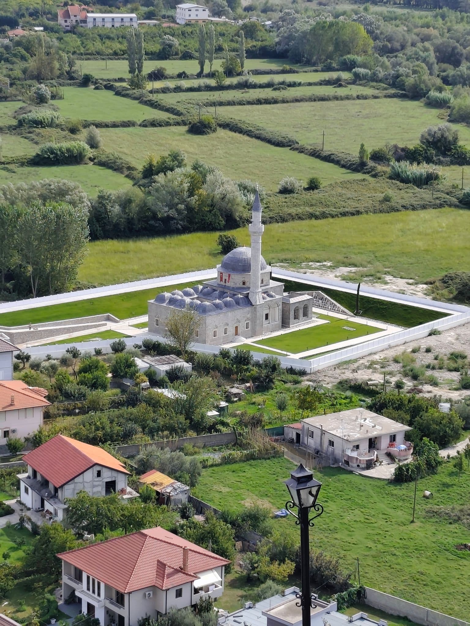 Stone Ottoman-style mosque with a single minaret seen from above, surrounded by green fields and nearby houses, Turkey.