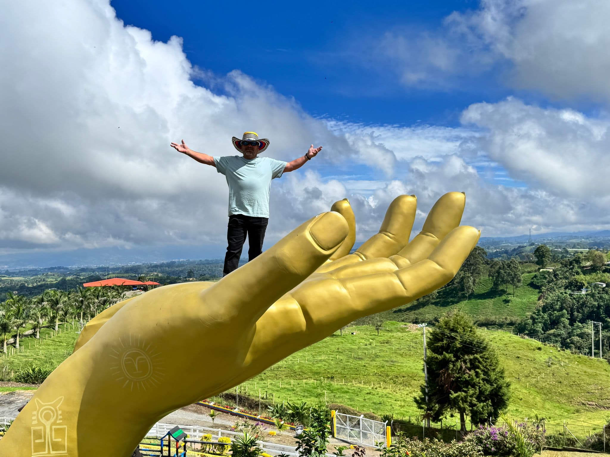 Large gold hand sculpture holding a person standing with arms outstretched at a hillside viewpoint in Filandia, Colombia.