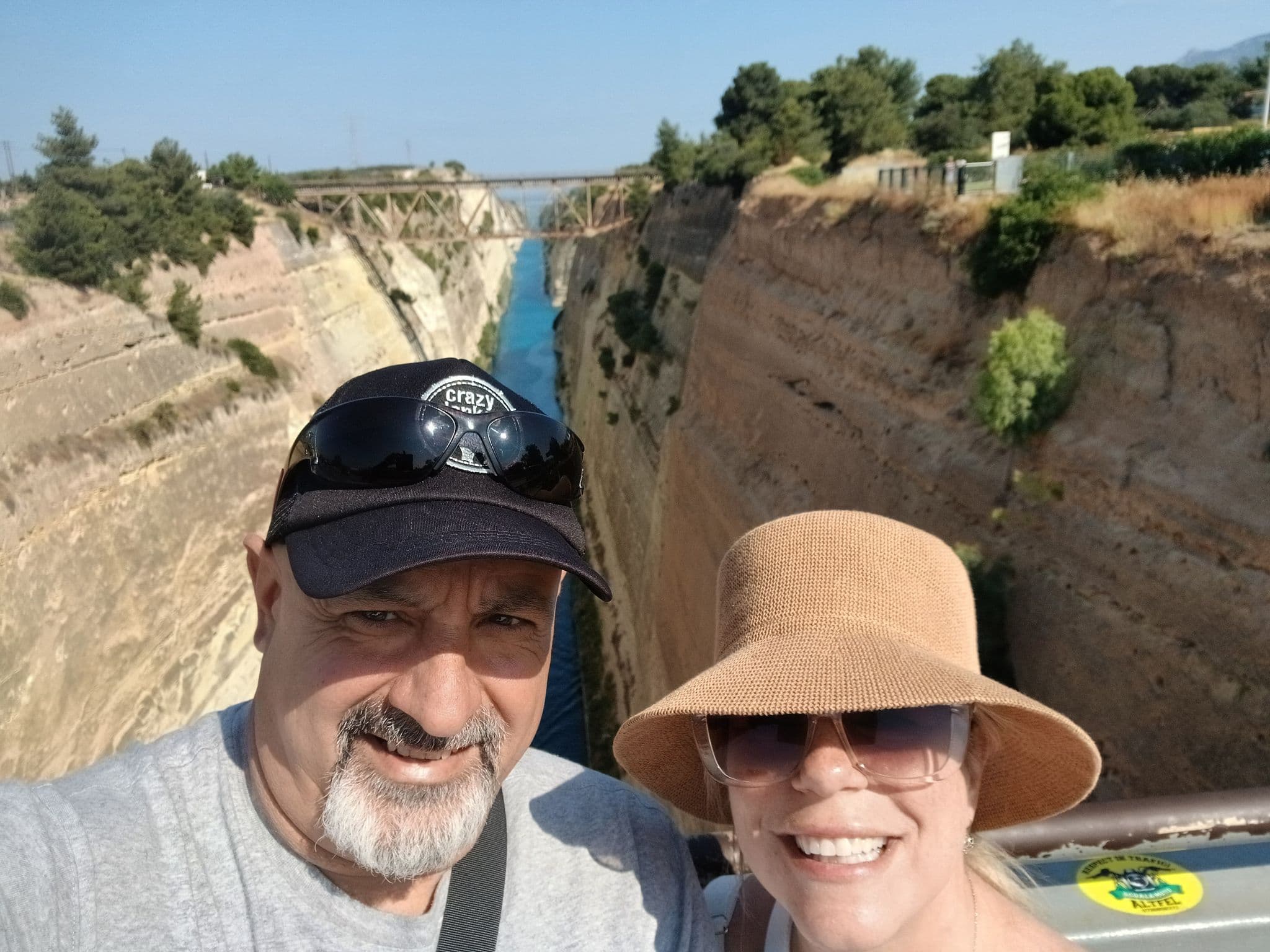 Corinth Canal seen from a bridge with two travelers taking a selfie on a sunny day, near Corinth, Greece.