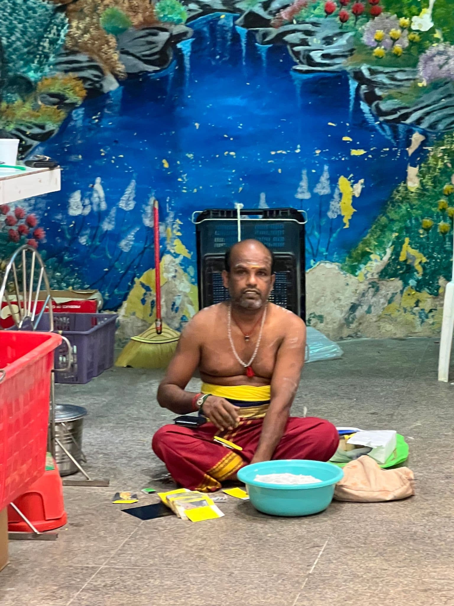A shirtless man sitting cross-legged sorting items beside a blue basin inside a decorated temple area at Batu Caves, Malaysia.