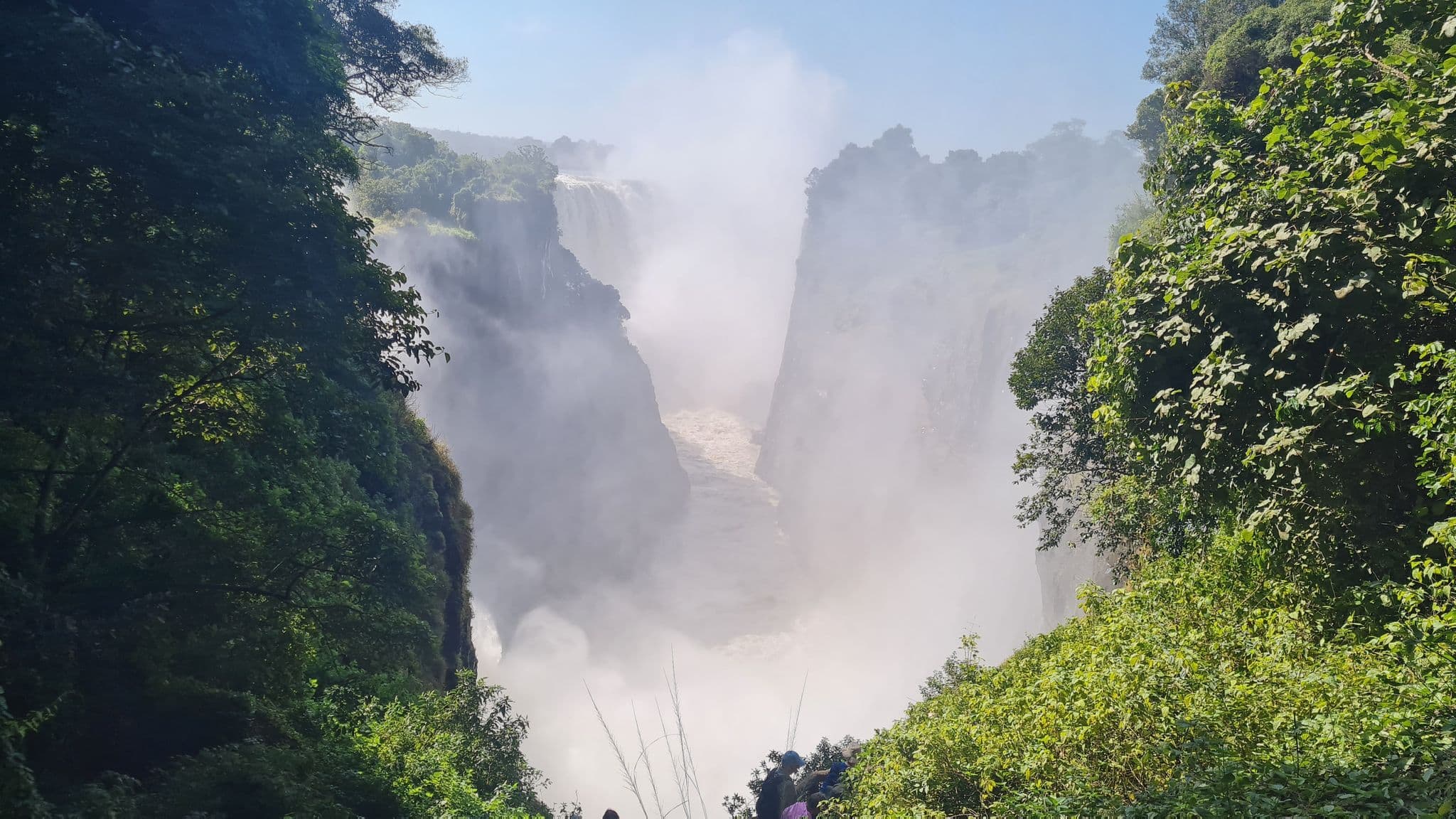Victoria Falls plunging into a mist-filled gorge, framed by lush green vegetation with tiny visitors visible on a nearby viewpoint.