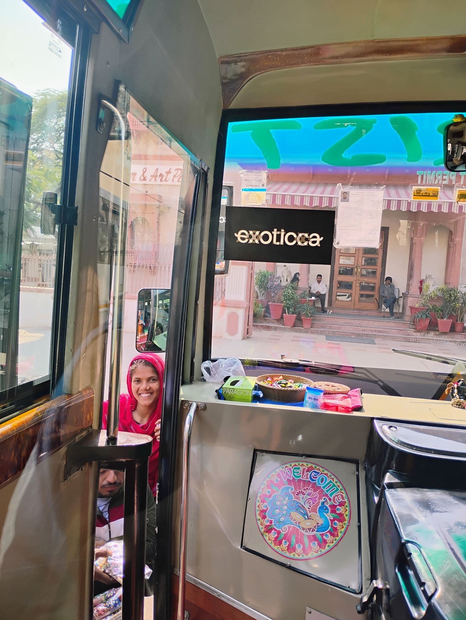 Smiling woman selling souvenirs at the open doorway of a tour bus, pink colonial-style building visible outside in India.