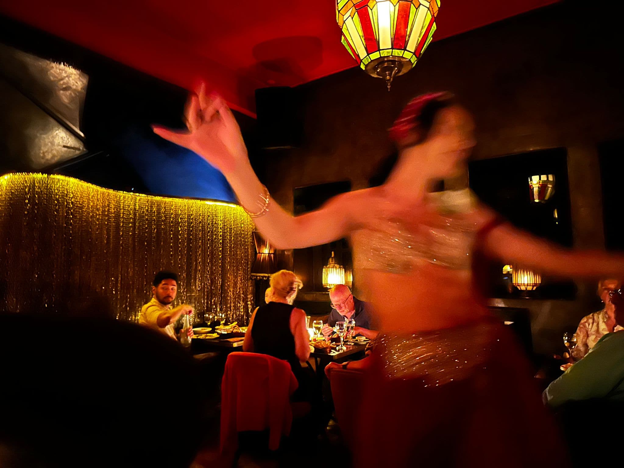Belly dancer captured mid-motion performing in a dim Marrakech restaurant while diners eat at nearby tables, Morocco.