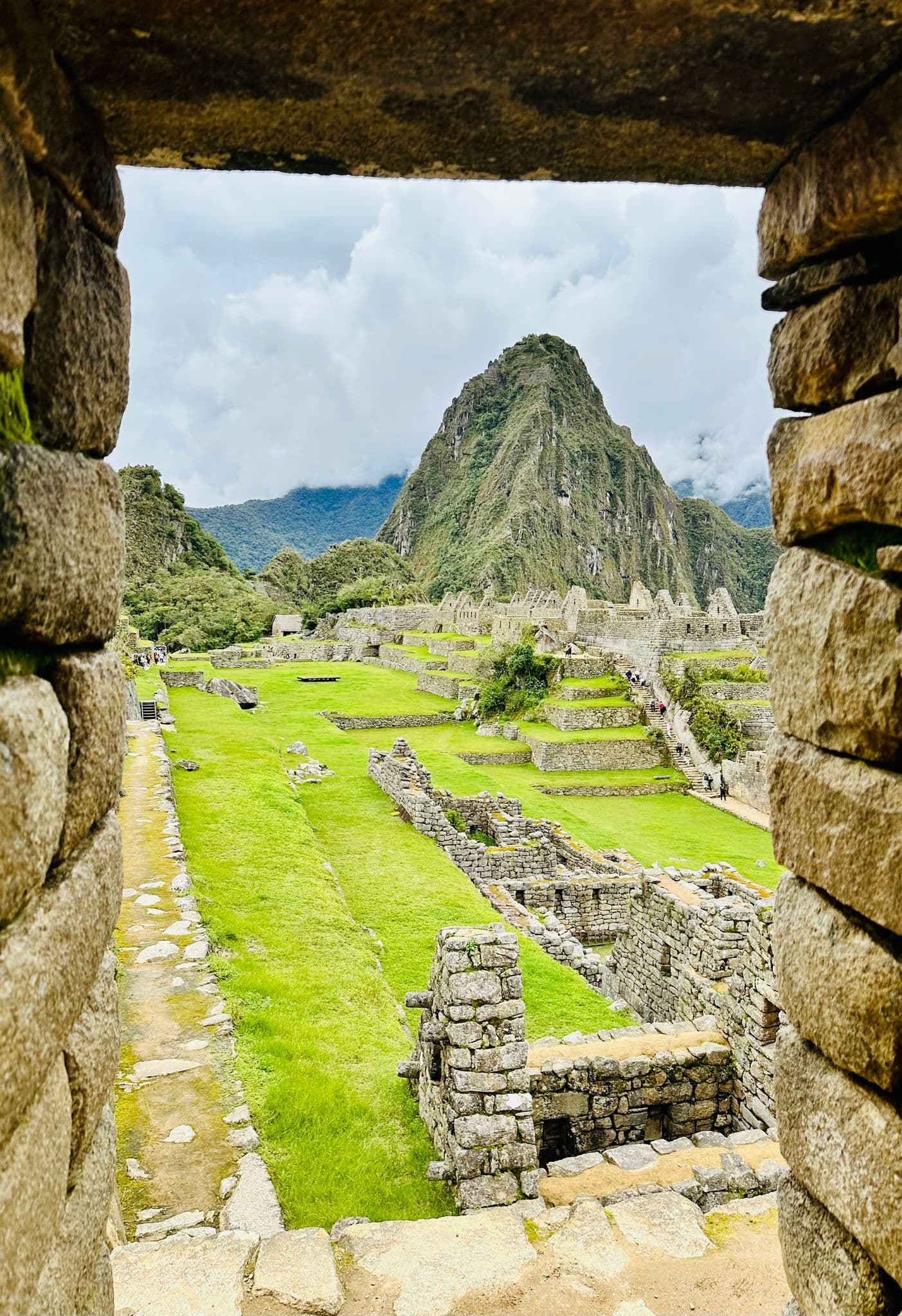 Machu Picchu ruins and Huayna Picchu framed through a stone window, terraces and small visitors visible in the Cusco region, Peru