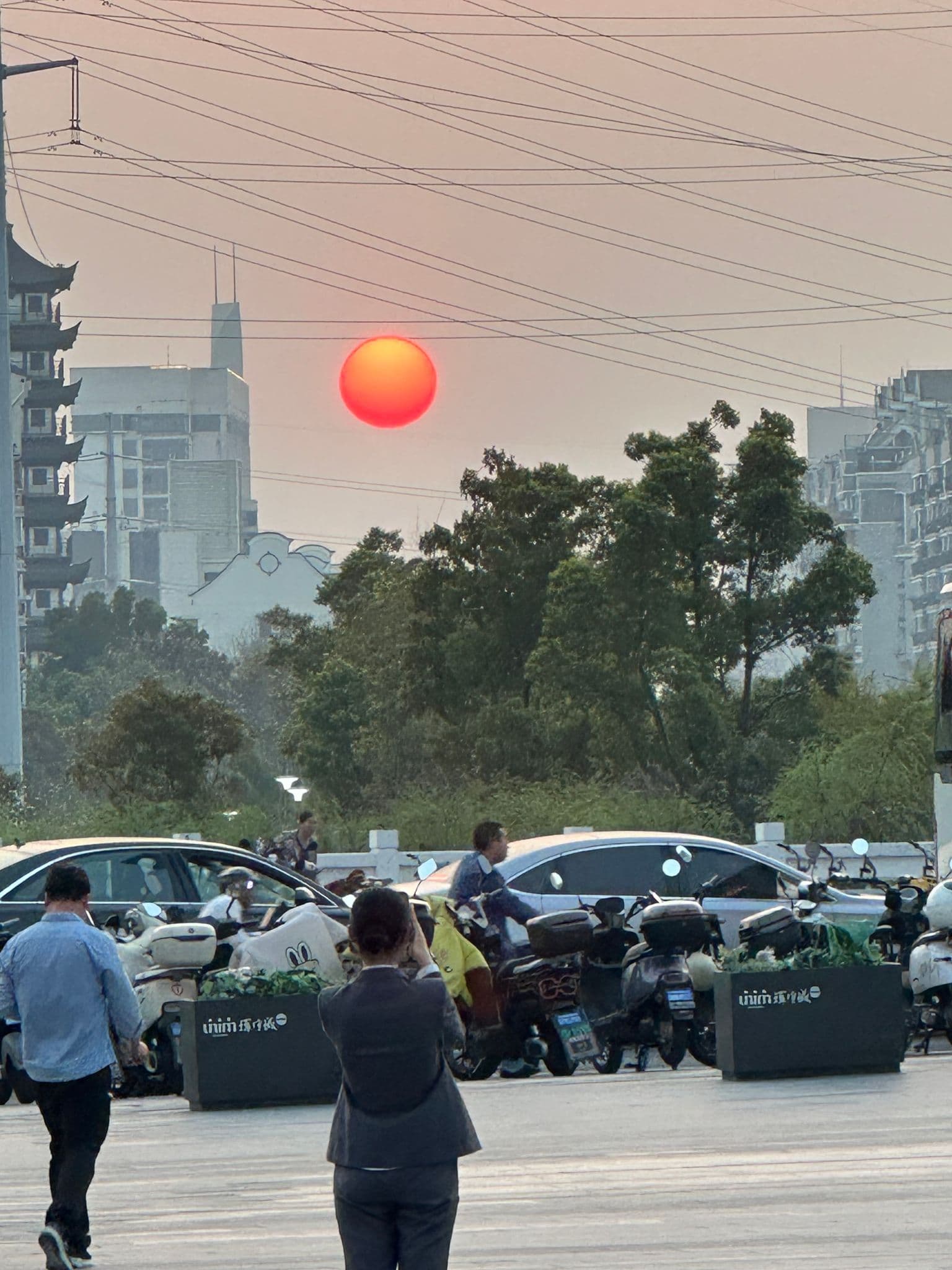 Red setting sun over an urban street with parked scooters and people taking photos, Shanghai, China.