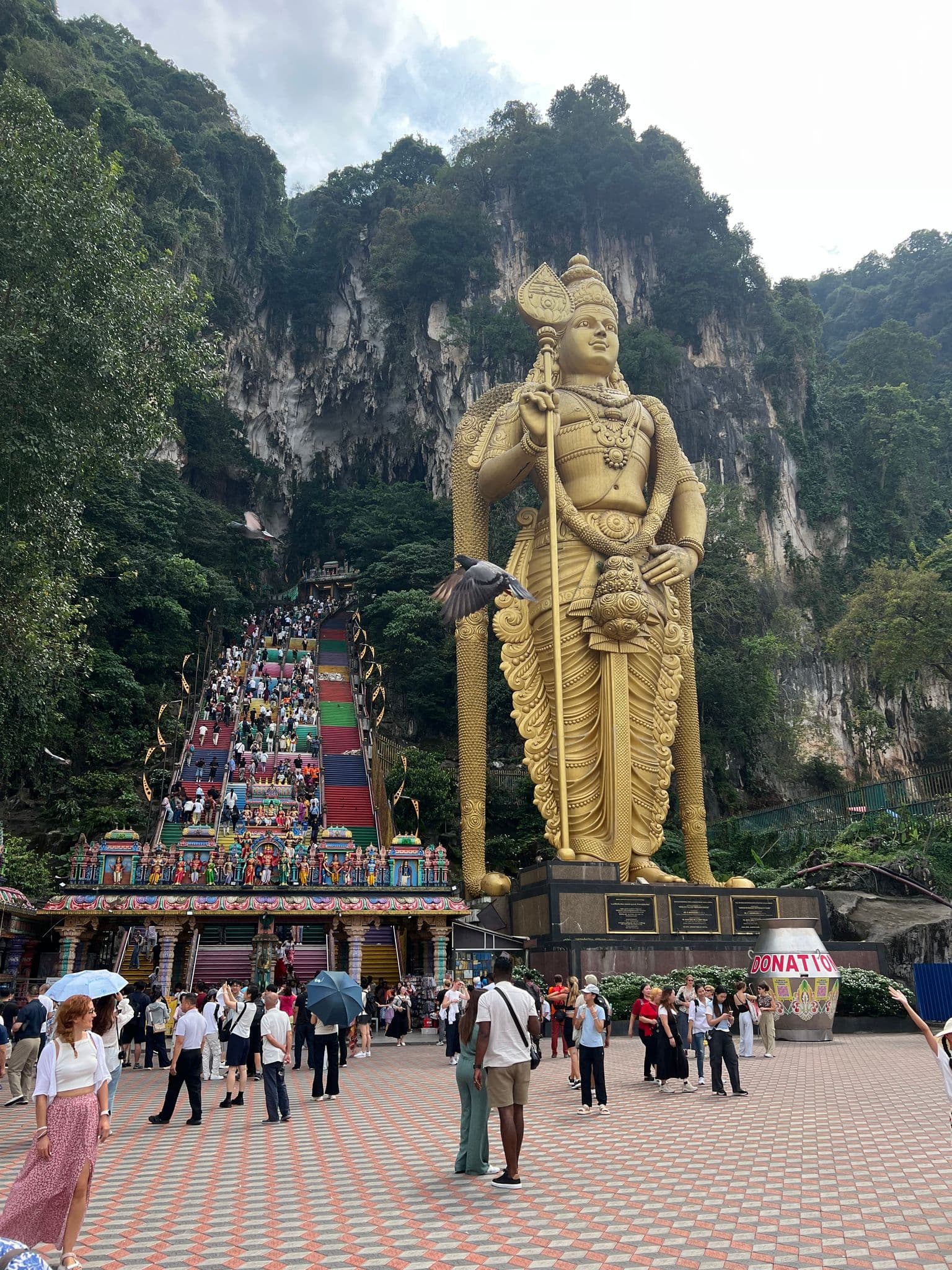 Golden Lord Murugan statue with the colorful staircase to Batu Caves in Kuala Lumpur, Malaysia, visitors climbing and gathering in the plaza.
