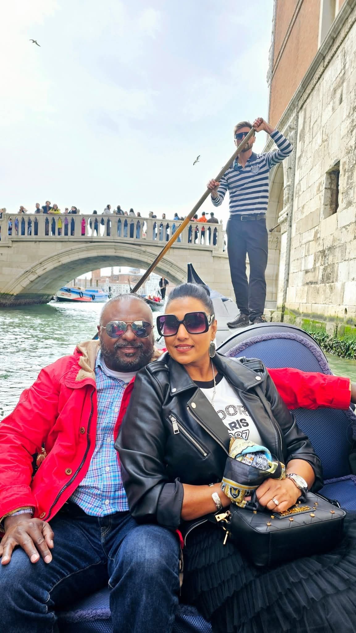 Couple seated in a gondola on a Venice canal while a gondolier stands and poles past a pedestrian bridge in Venice, Italy.