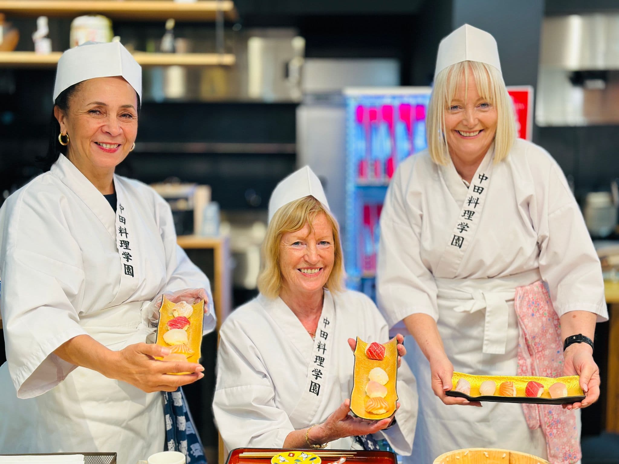 Sushi-making class with three women in chef robes holding plated sushi, Japan.