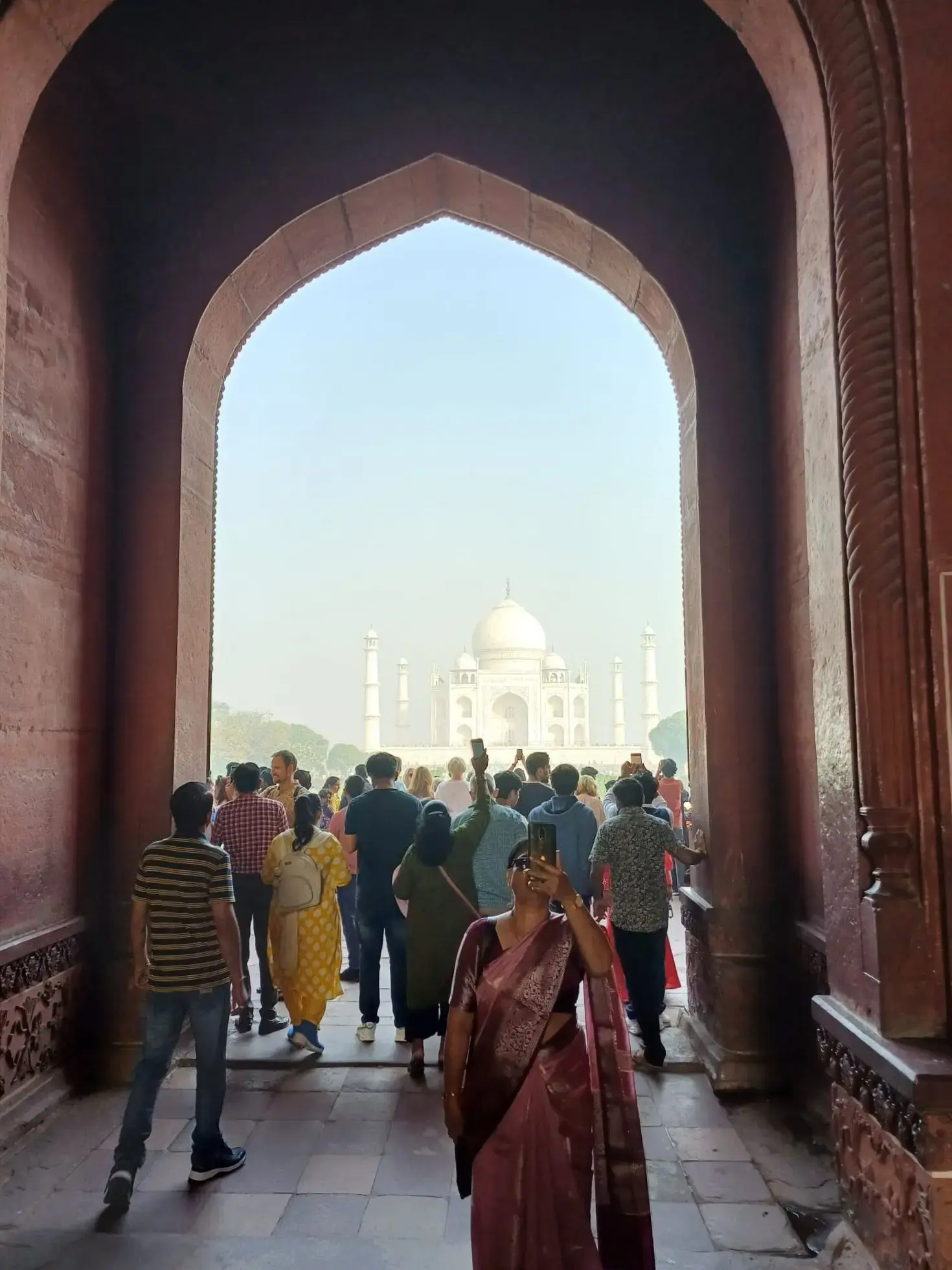Taj Mahal framed by a red sandstone archway, crowds approaching it while a woman in a pink sari takes a selfie, Agra, India.
