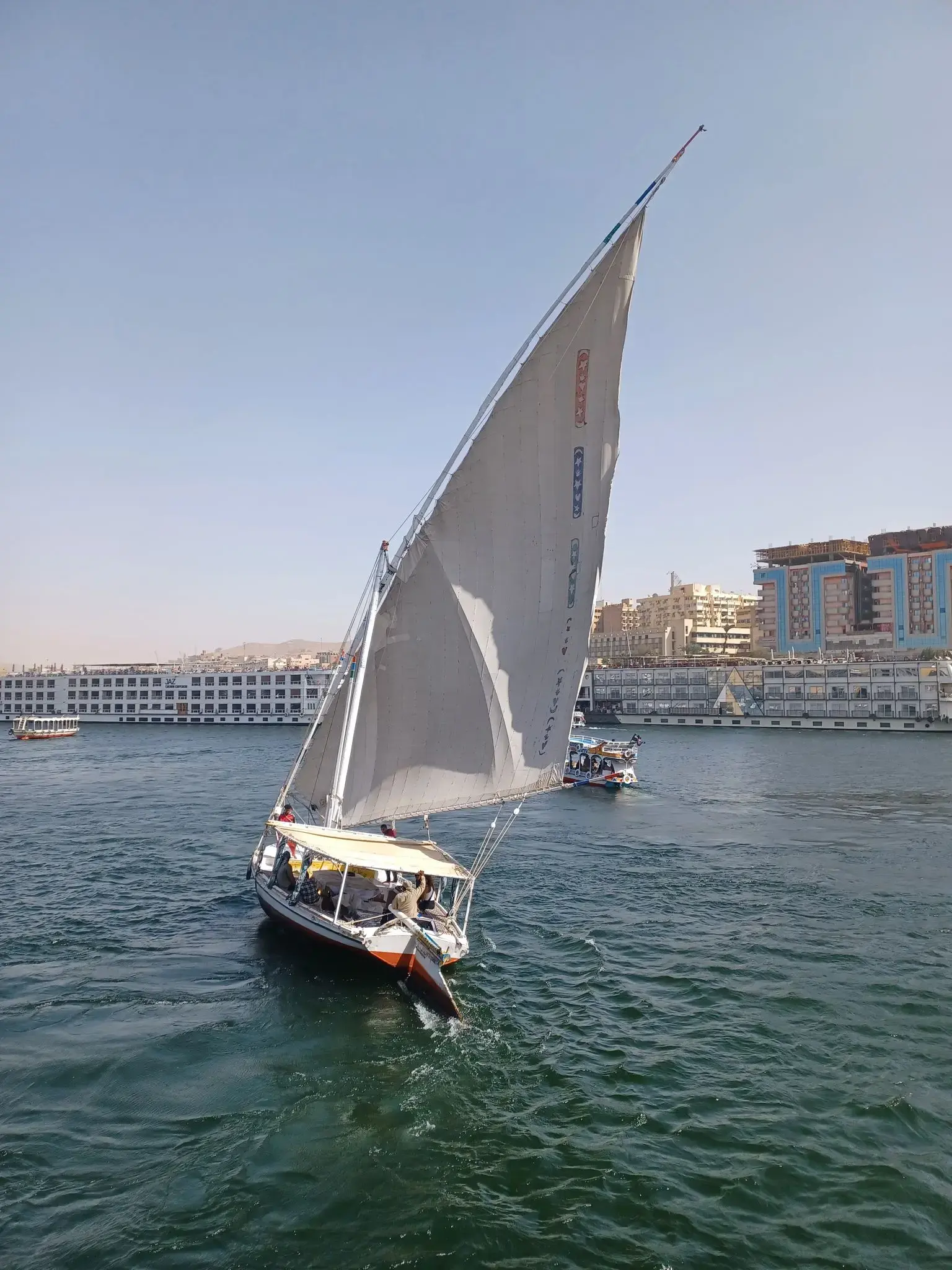Felucca sailboat on the Nile River with passengers sailing past riverfront buildings in Egypt.