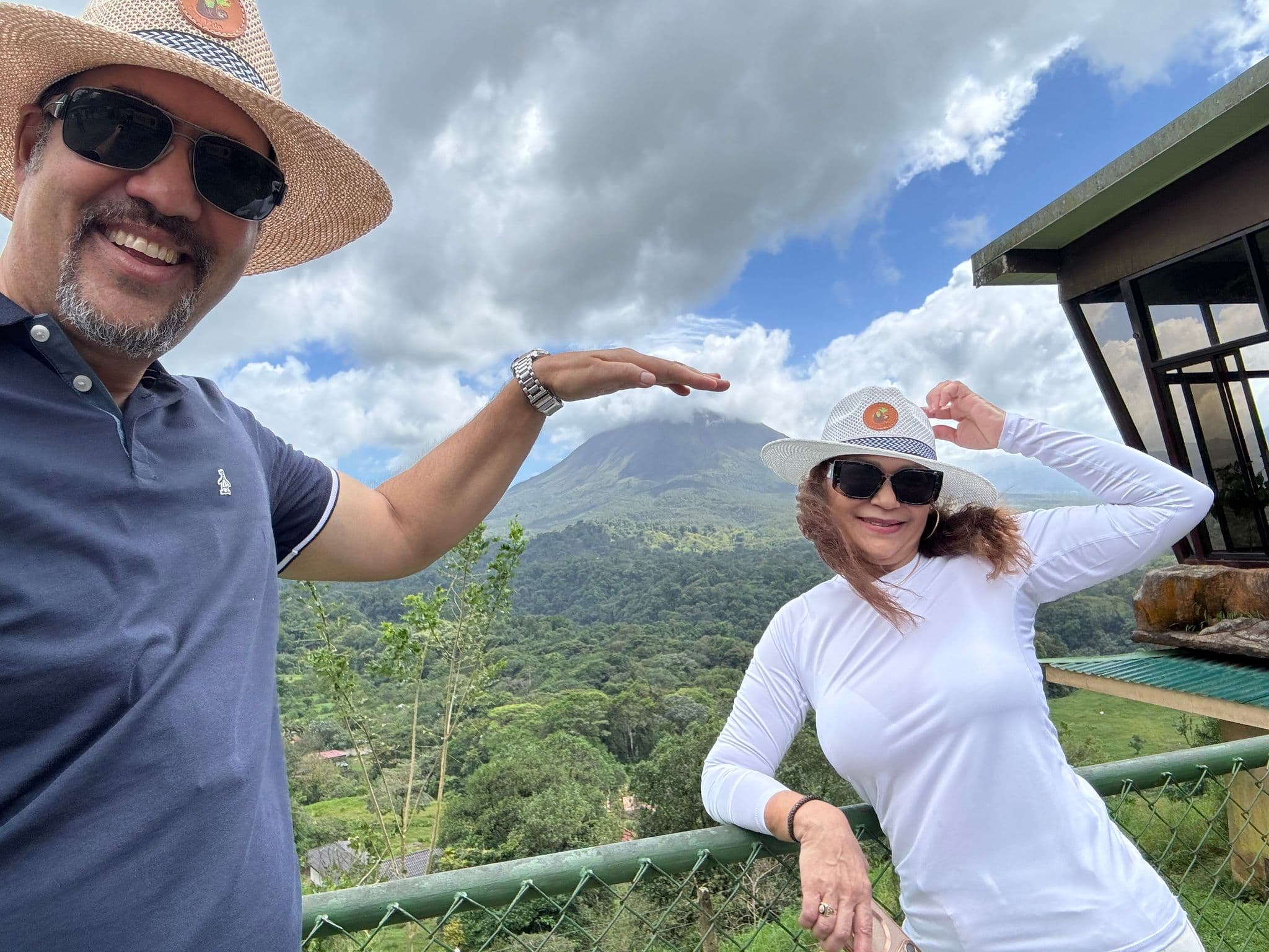 Arenal Volcano with two travelers posing at a viewpoint, the man framing the peak with his hand, Costa Rica.