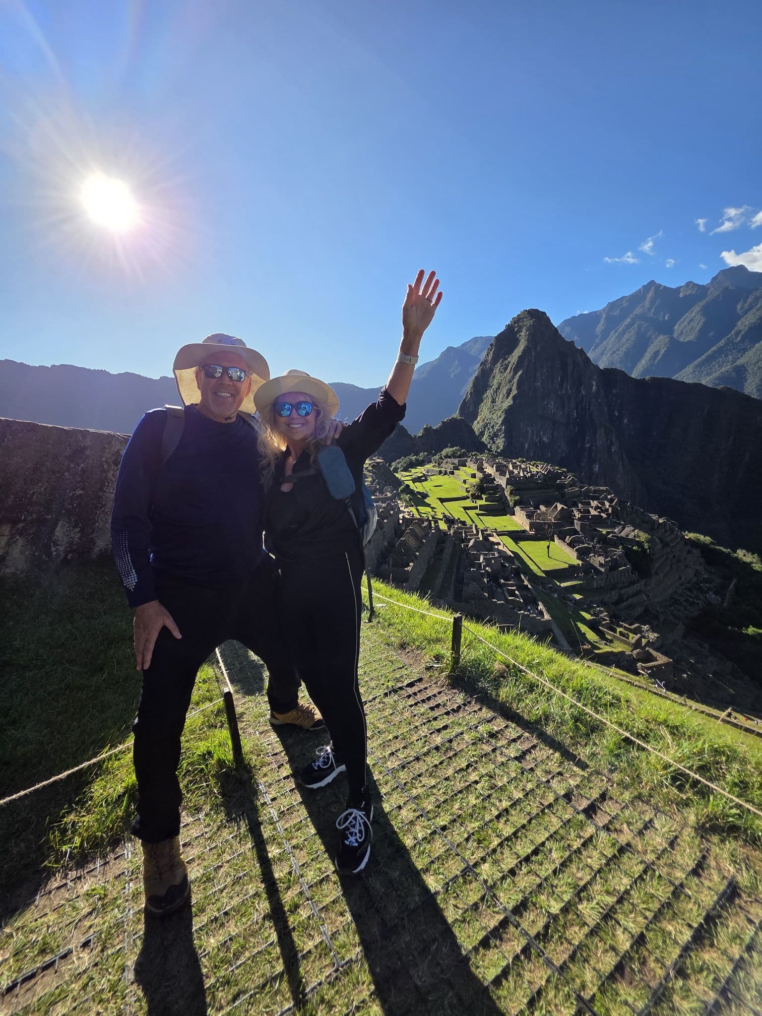 Machu Picchu ruins with Huayna Picchu in the background and two travelers posing on a grassy overlook, Cusco region, Peru.
