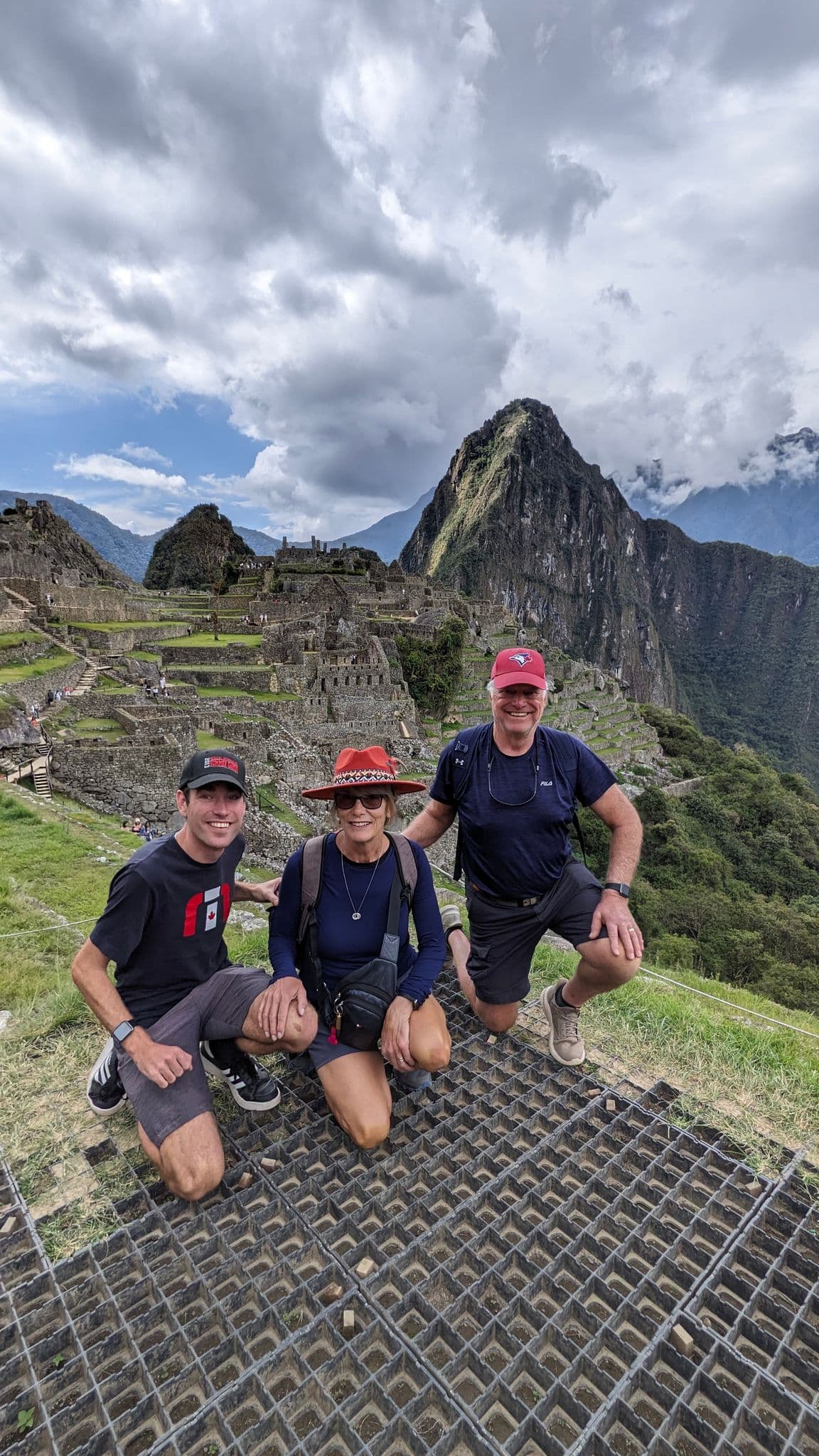 Machu Picchu citadel with three travelers kneeling on a grassy terrace and Huayna Picchu peak in the background, Cusco Region, Peru.