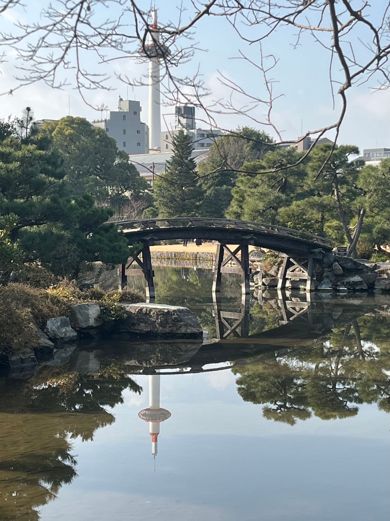 Kyoto Tower reflected in a calm pond with a wooden arched bridge and trees in a Kyoto park, Japan.