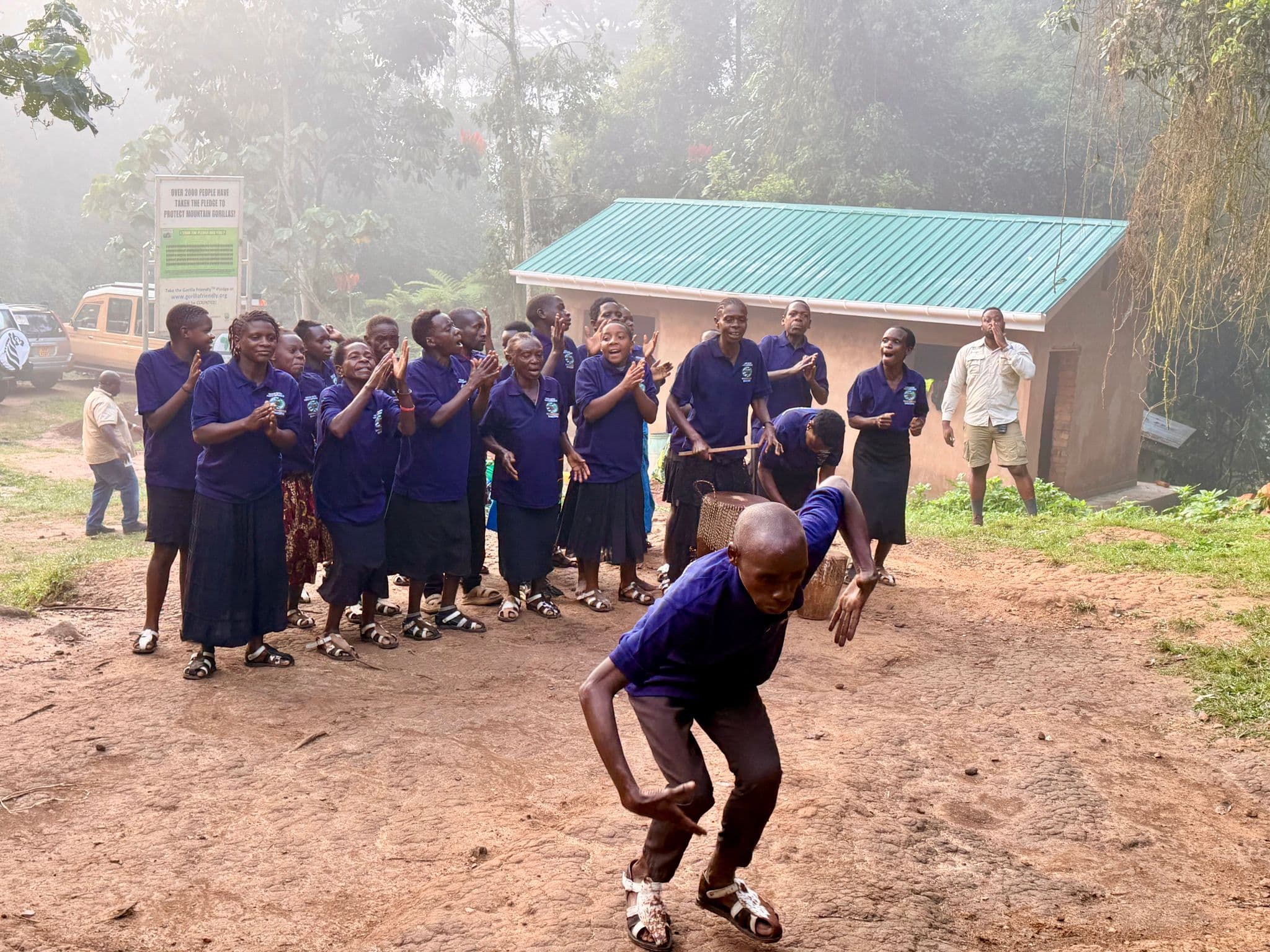 A dancer in blue leaning forward while a group of clapping performers stand outside a small building in Bwindi Impenetrable Forest, Uganda.