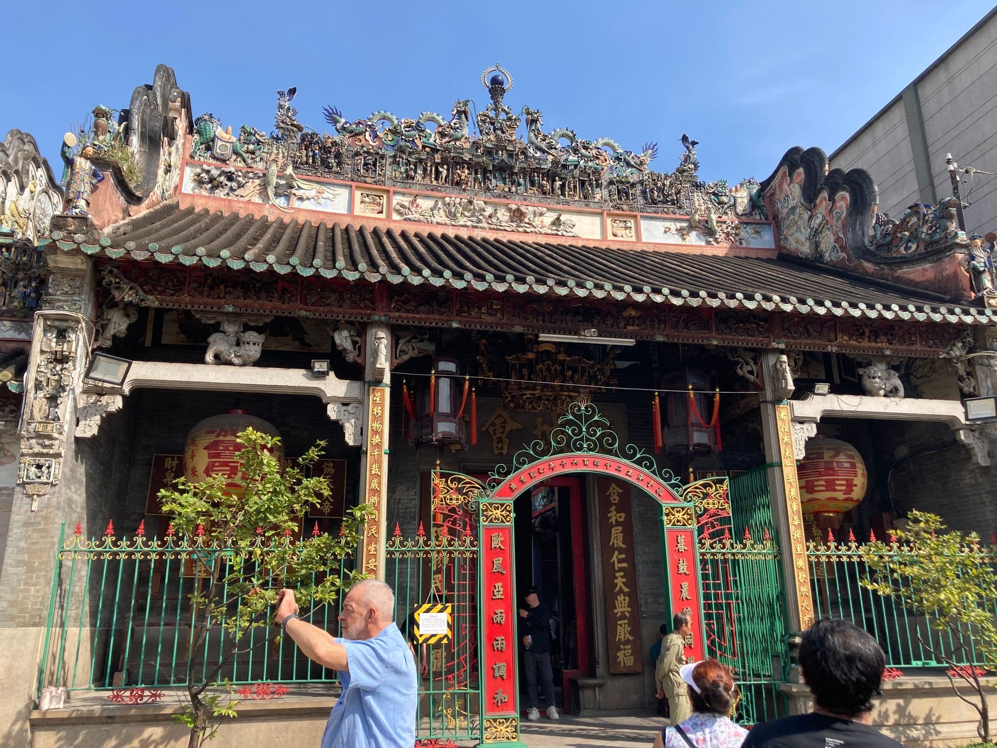Ornate Chinese temple facade with decorative roof and red lanterns, visitors entering and a man taking a photo.