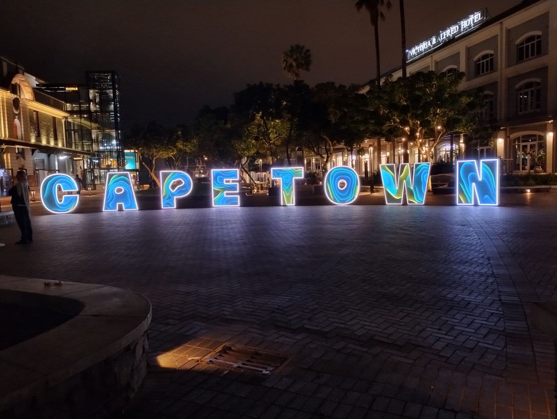 Illuminated "CAPE TOWN" sign at the V&A Waterfront, Cape Town, South Africa, lit at night with a few people nearby.