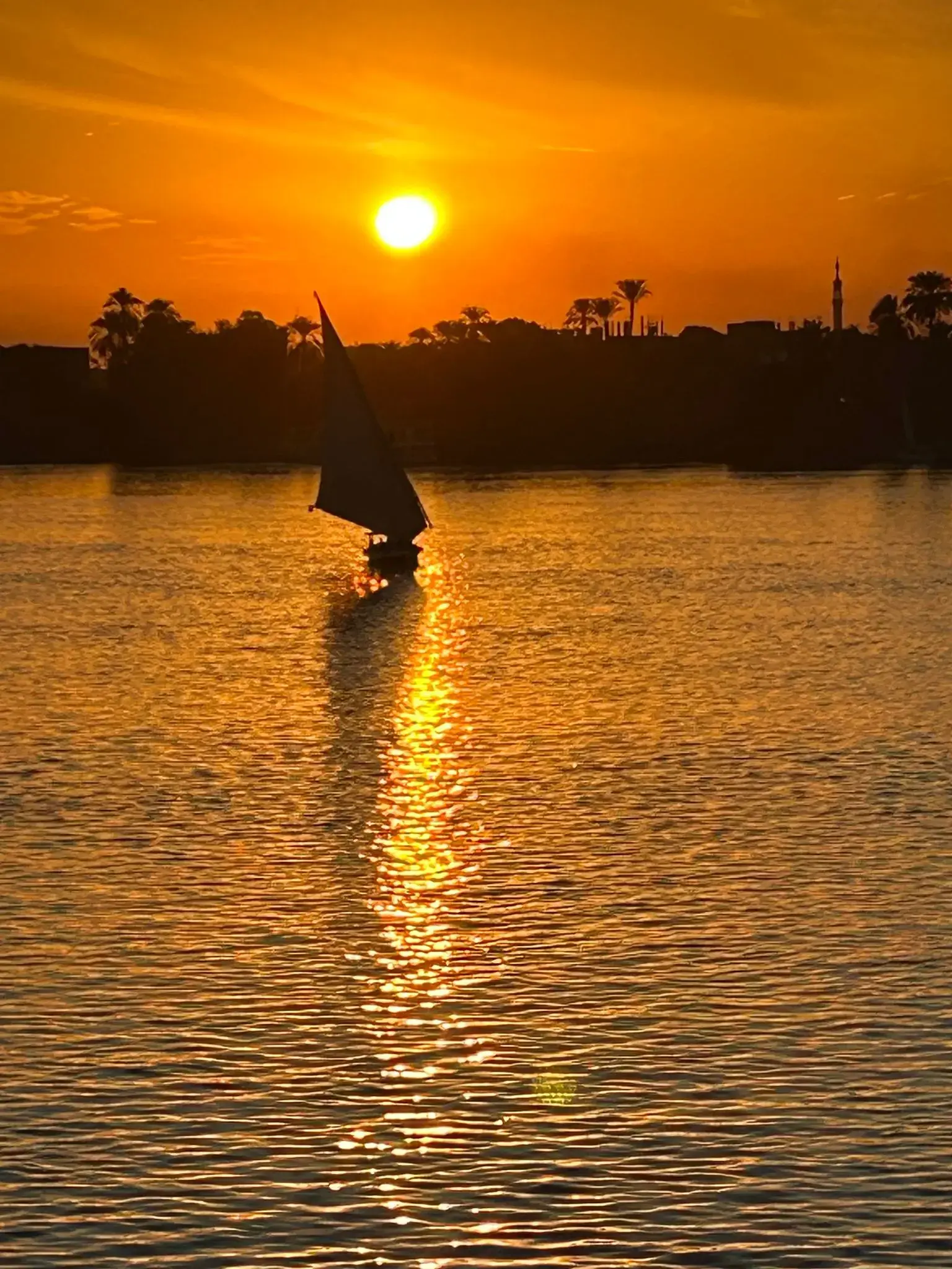 Nile River at sunset with a felucca sailing, the sun reflecting on the water and a silhouetted shoreline in Egypt.