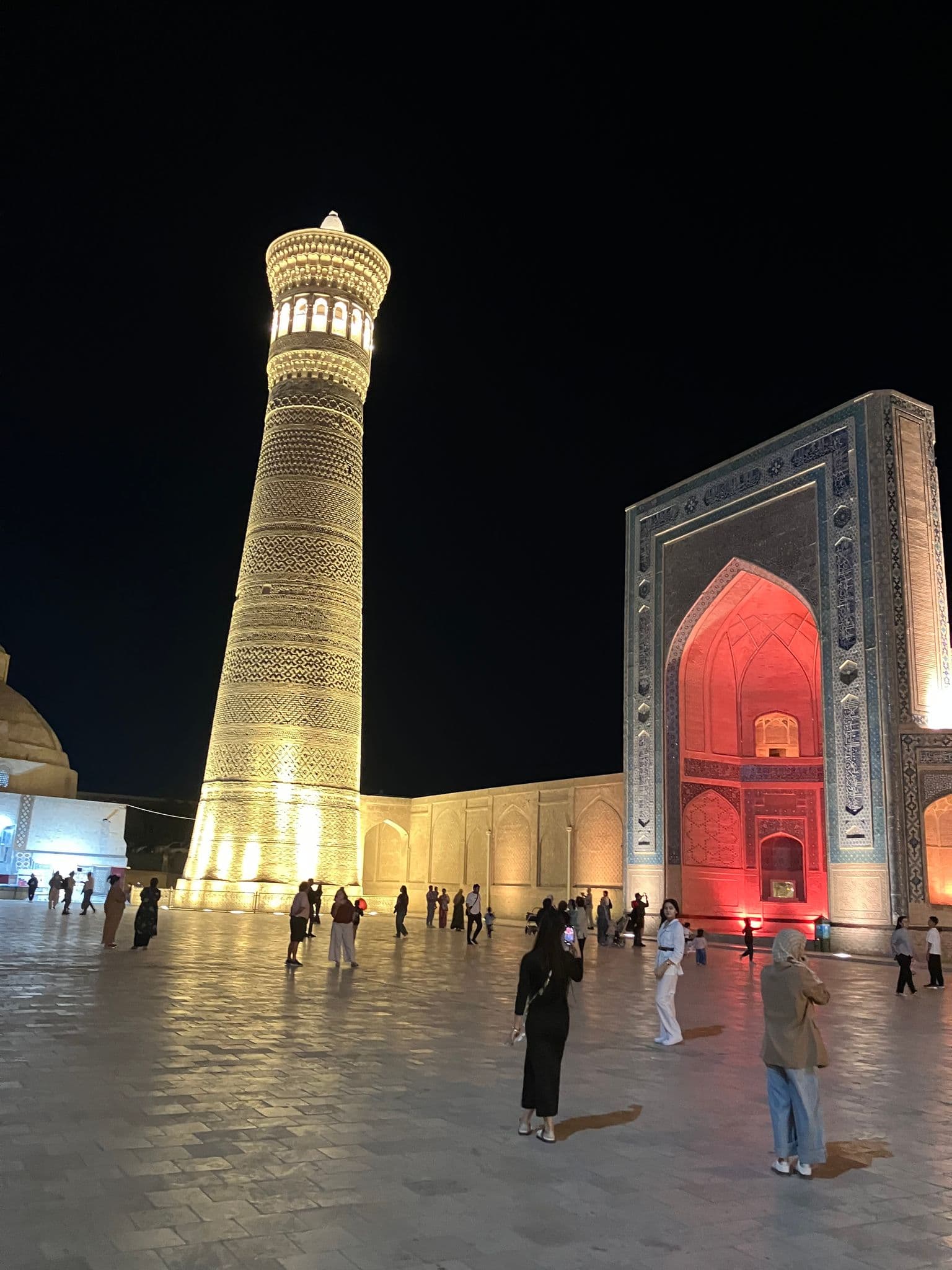 Illuminated Kalyan Minaret at Po-i-Kalyan in Bukhara, Uzbekistan, with people walking and taking photos in the night plaza.