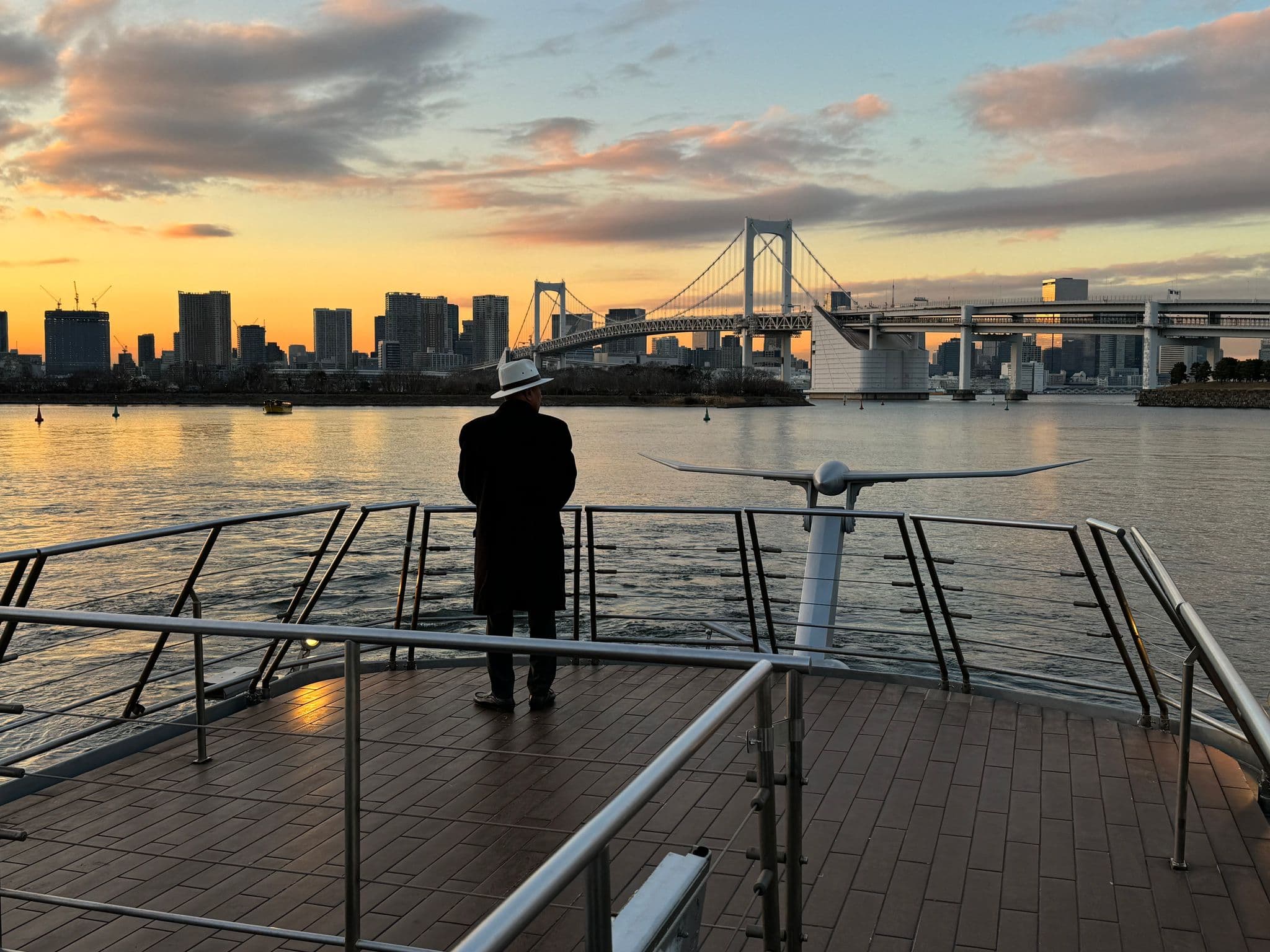 Rainbow Bridge over Tokyo Bay at sunset, a person stands on a boat deck looking toward the Odaiba skyline, Tokyo, Japan