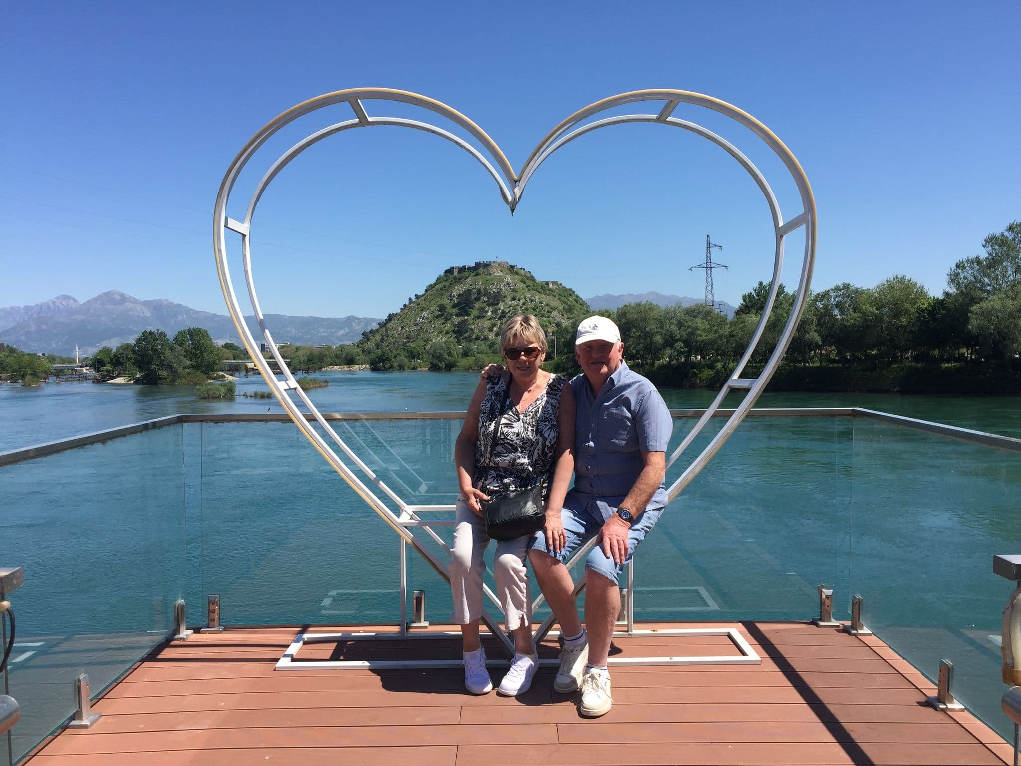 Heart-shaped metal frame with a couple sitting on a riverside viewing deck, a tree-covered hill and river behind them.
