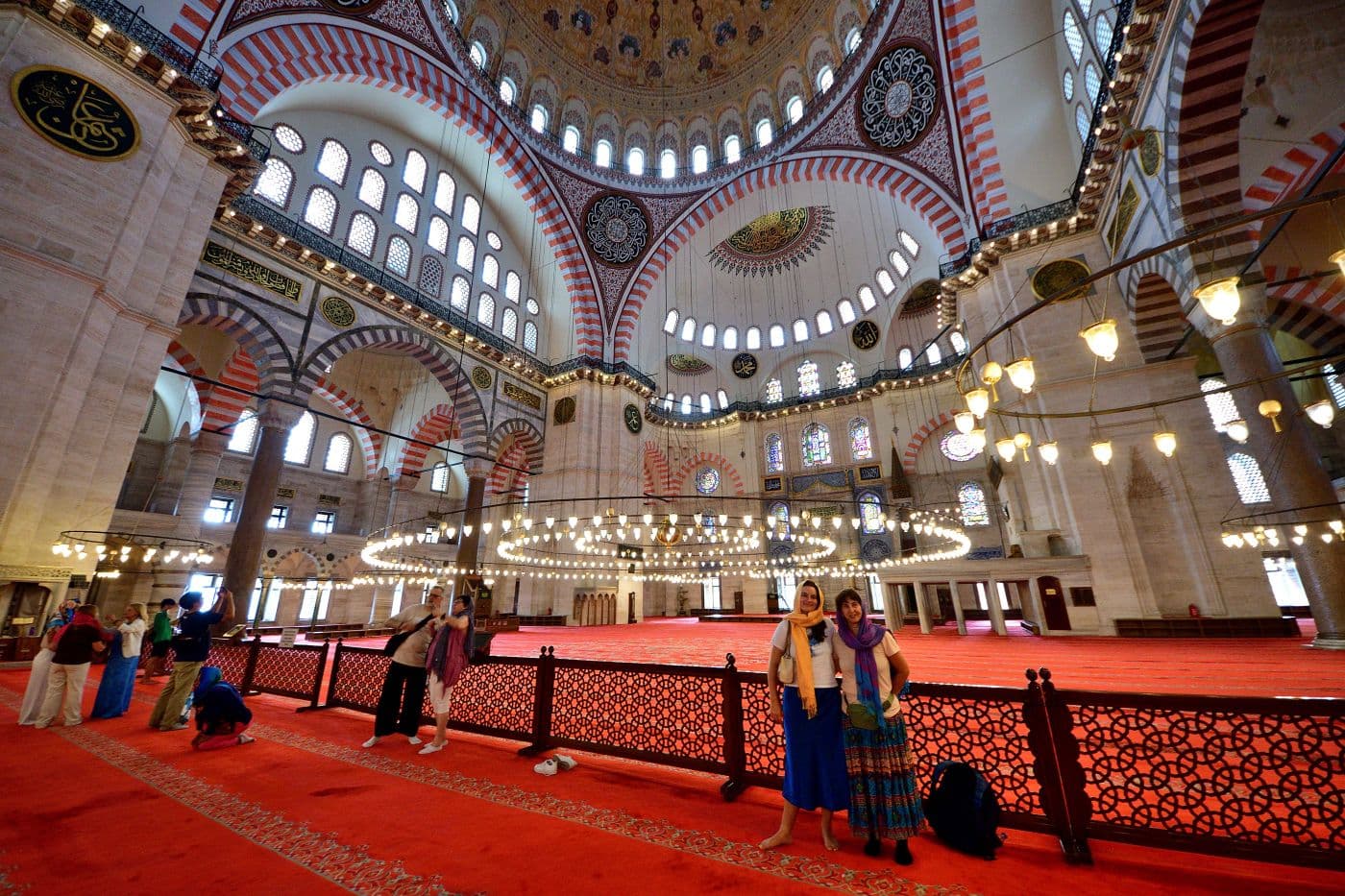 Interior of Süleymaniye Mosque in Istanbul with two travelers posing near the prayer hall and chandeliers above