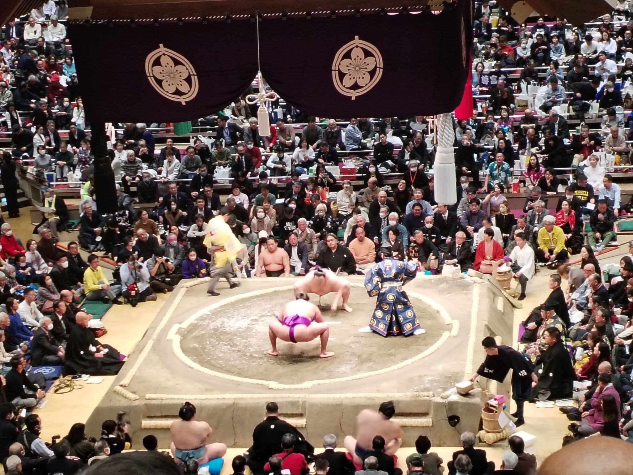 Sumo ring at Ryogoku Kokugikan with wrestlers facing off while a packed audience watches in Tokyo, Japan.