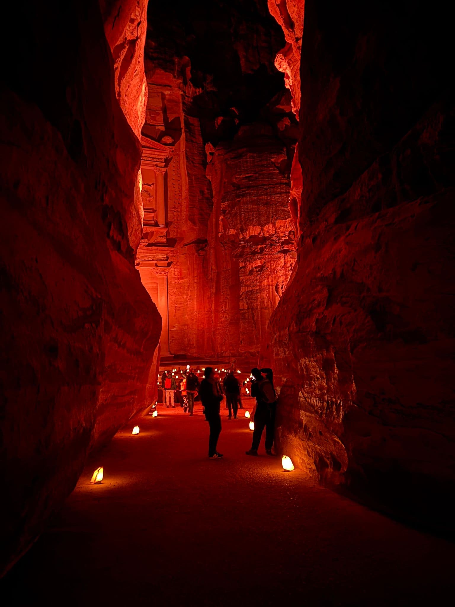 Al-Khazneh (The Treasury) at Petra lit red by lanterns, people walking and photographing in the narrow canyon, Jordan.