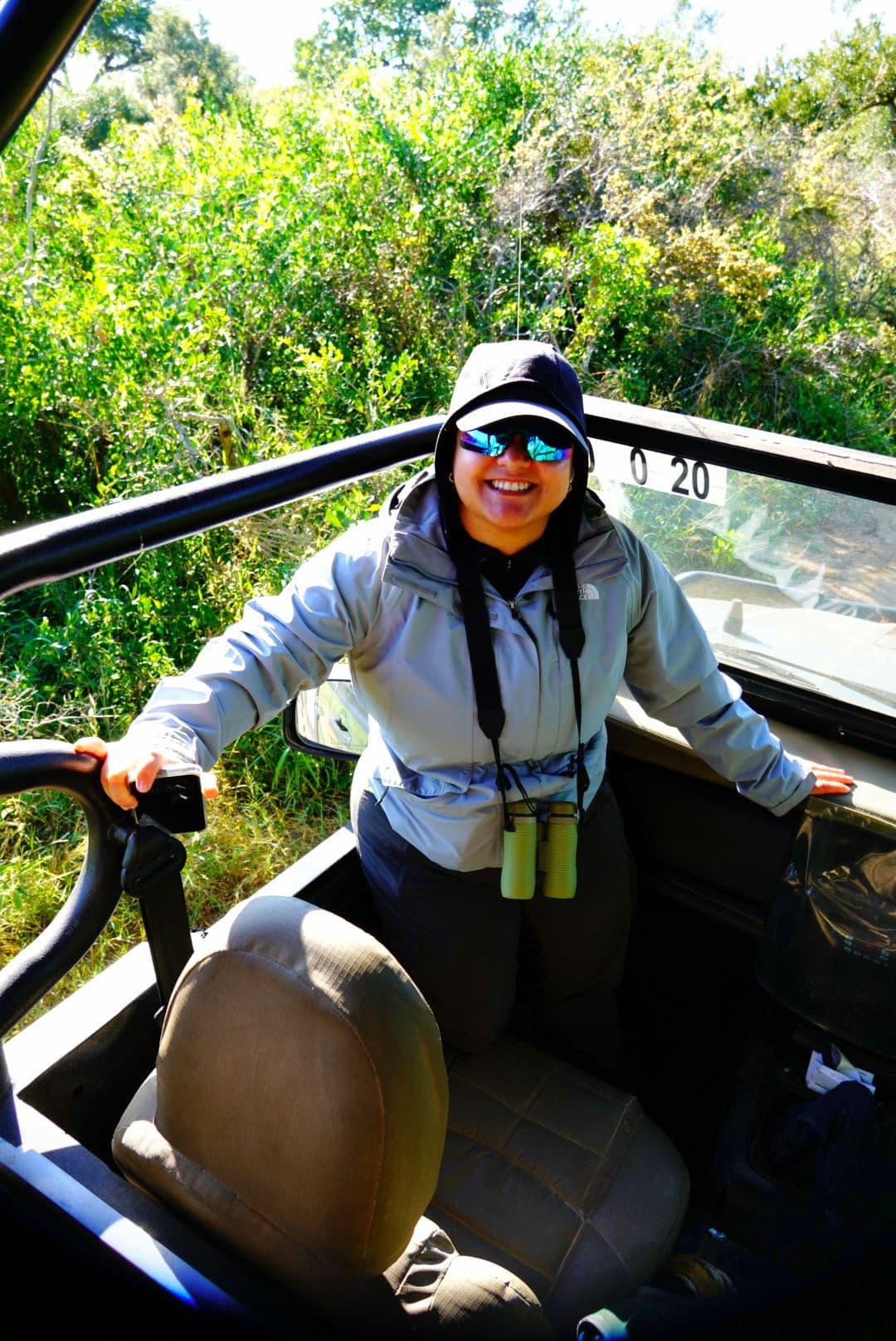 Smiling traveler standing in an open safari vehicle with binoculars around their neck in Kruger National Park, South Africa.