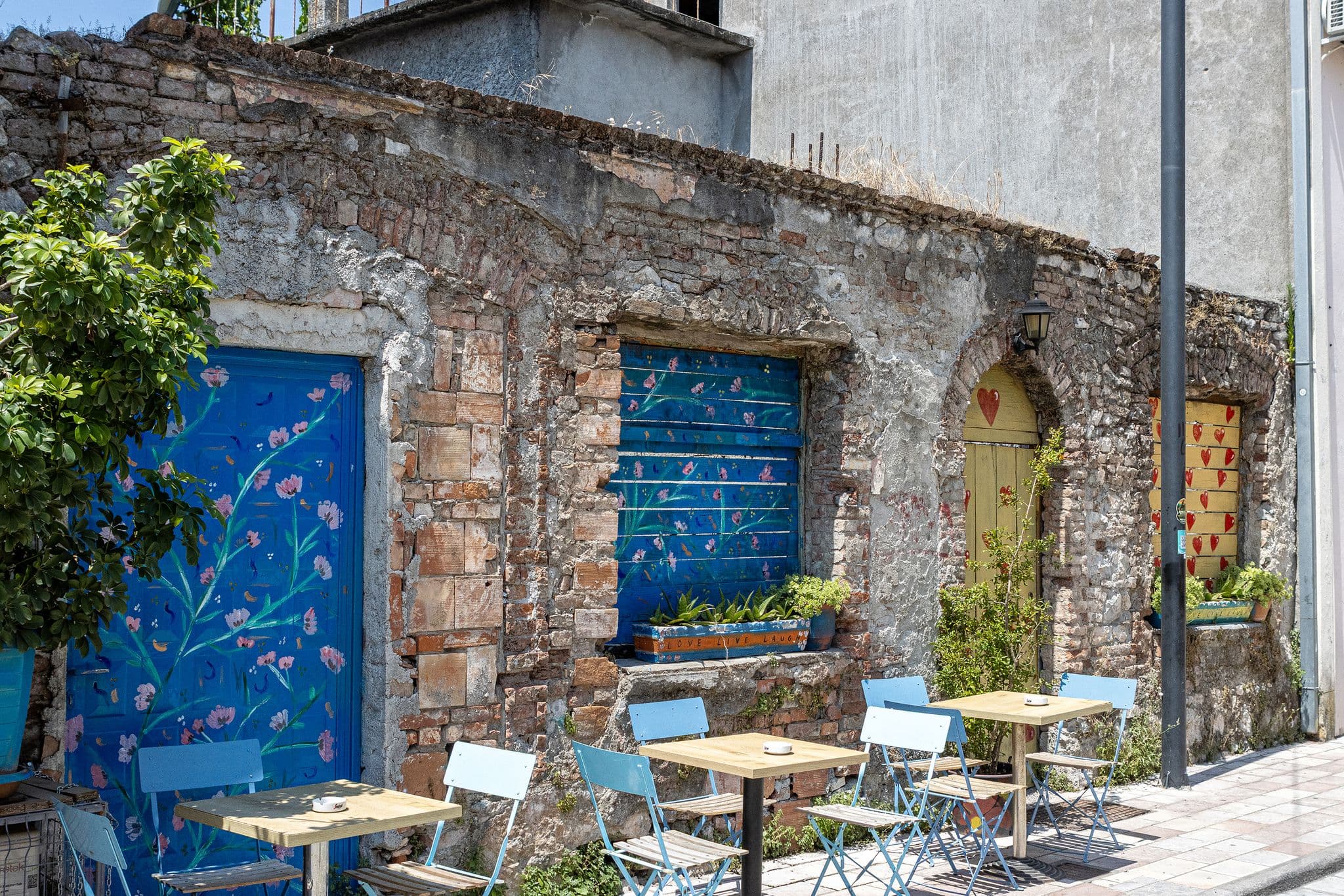 Painted wooden doors and shuttered windows on a weathered stone wall with empty blue cafe tables and chairs on a street in Shkodër, Albania.