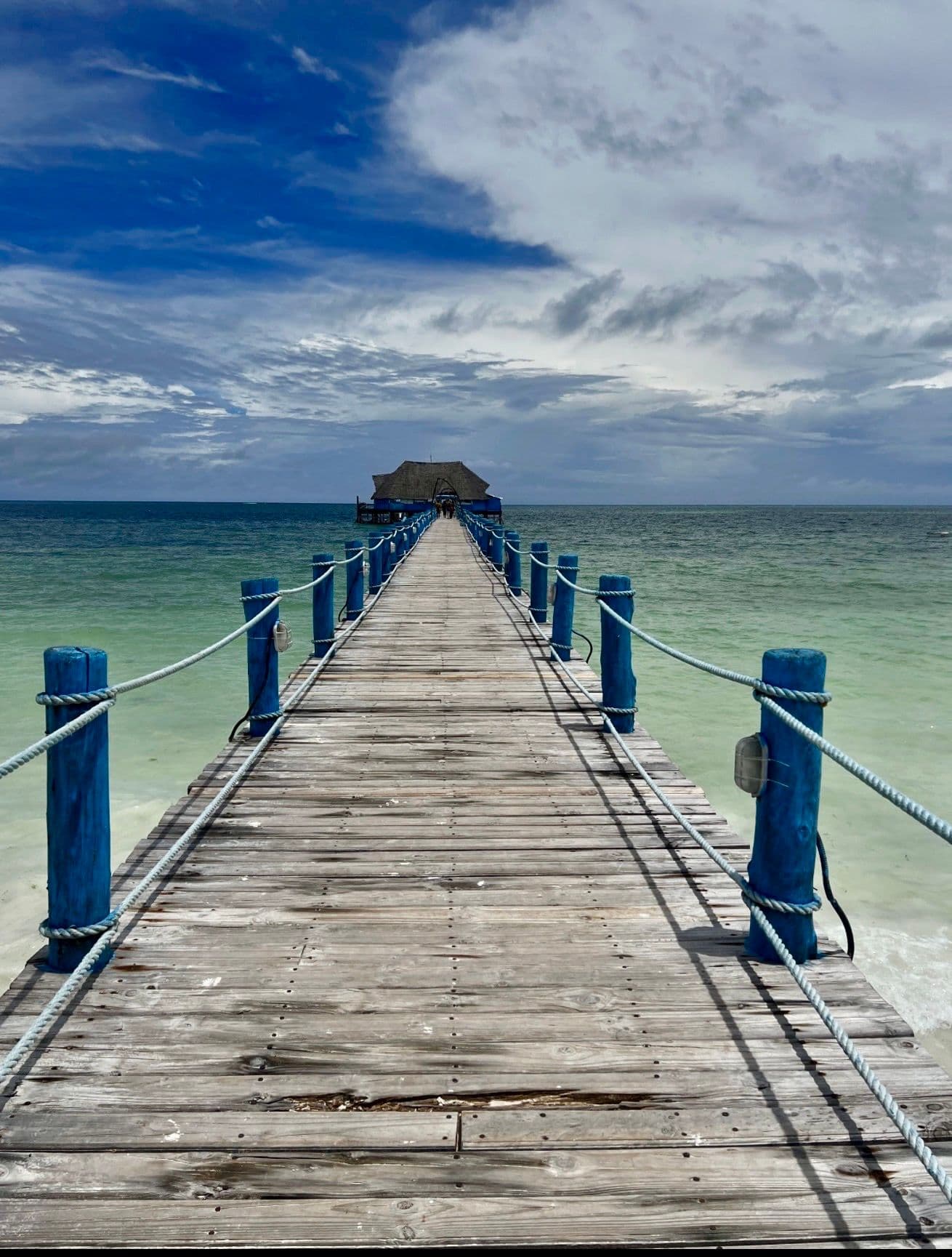 Wooden pier with blue posts stretching to a thatched jetty over turquoise water on Zanzibar, Tanzania.