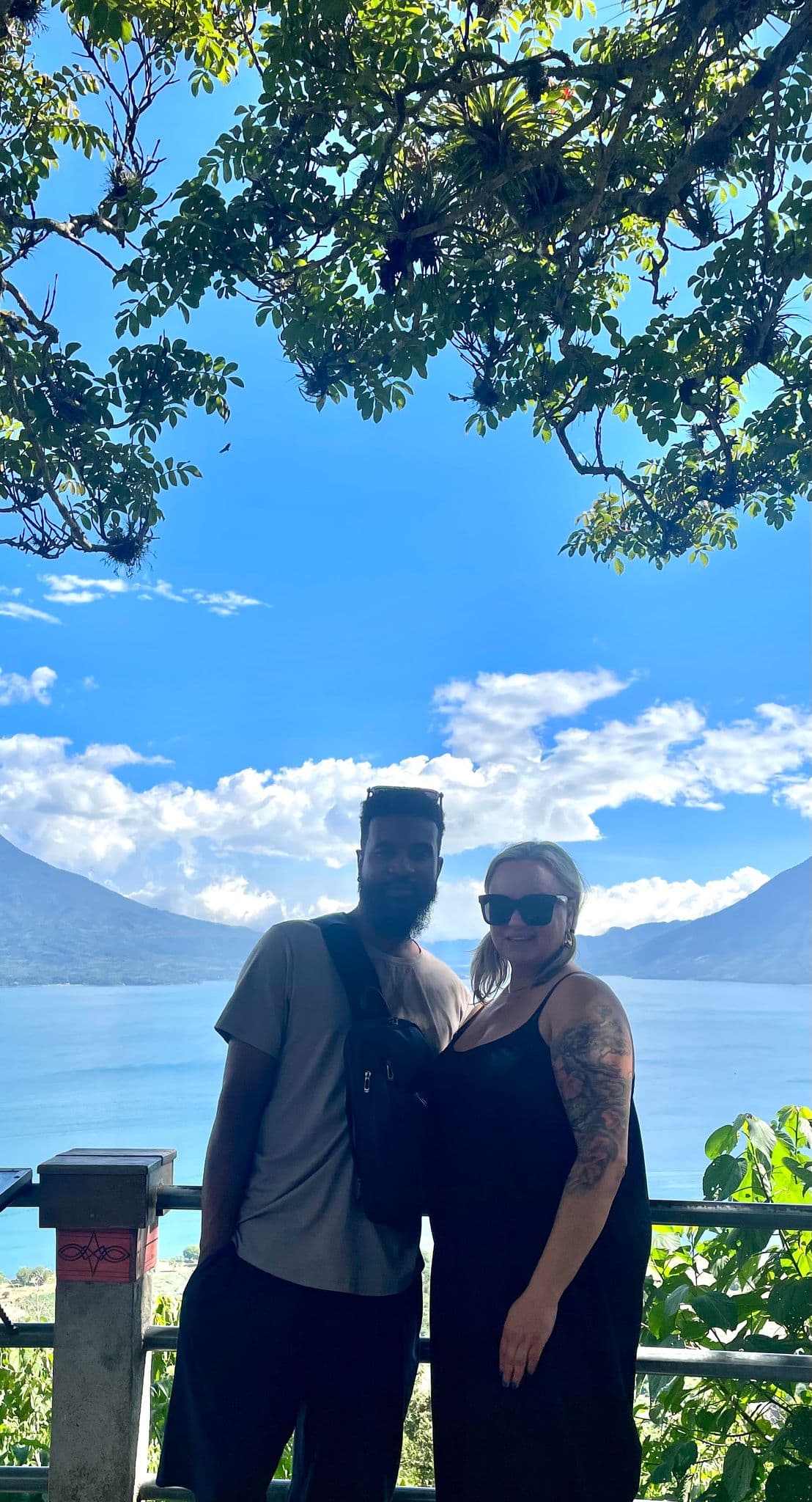 Lake Atitlán with volcano-lined shore behind two travelers posing at a hillside viewpoint, Guatemala.